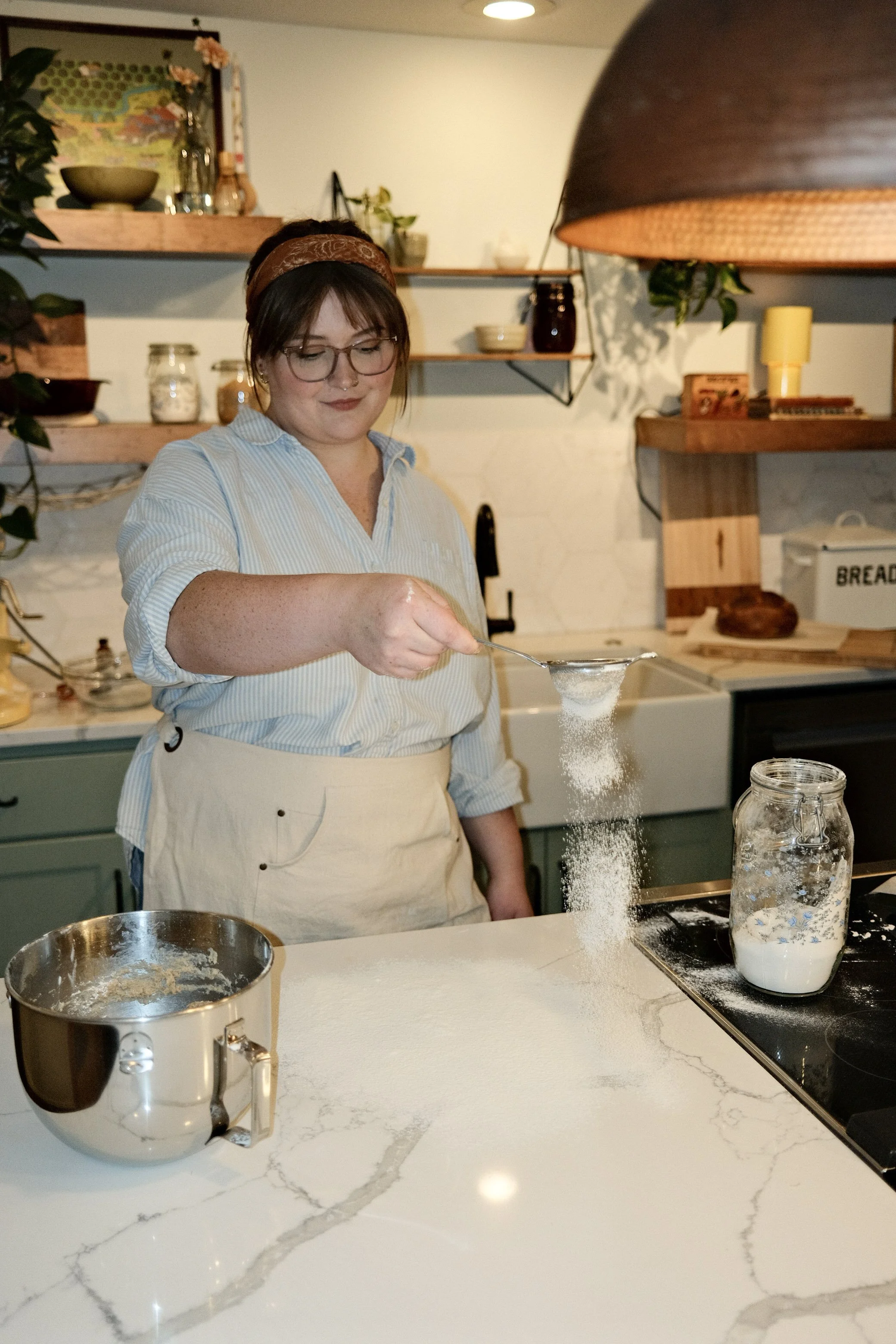 A woman with glasses and a headband sifting flour into a jar on a kitchen countertop with a white marble surface.