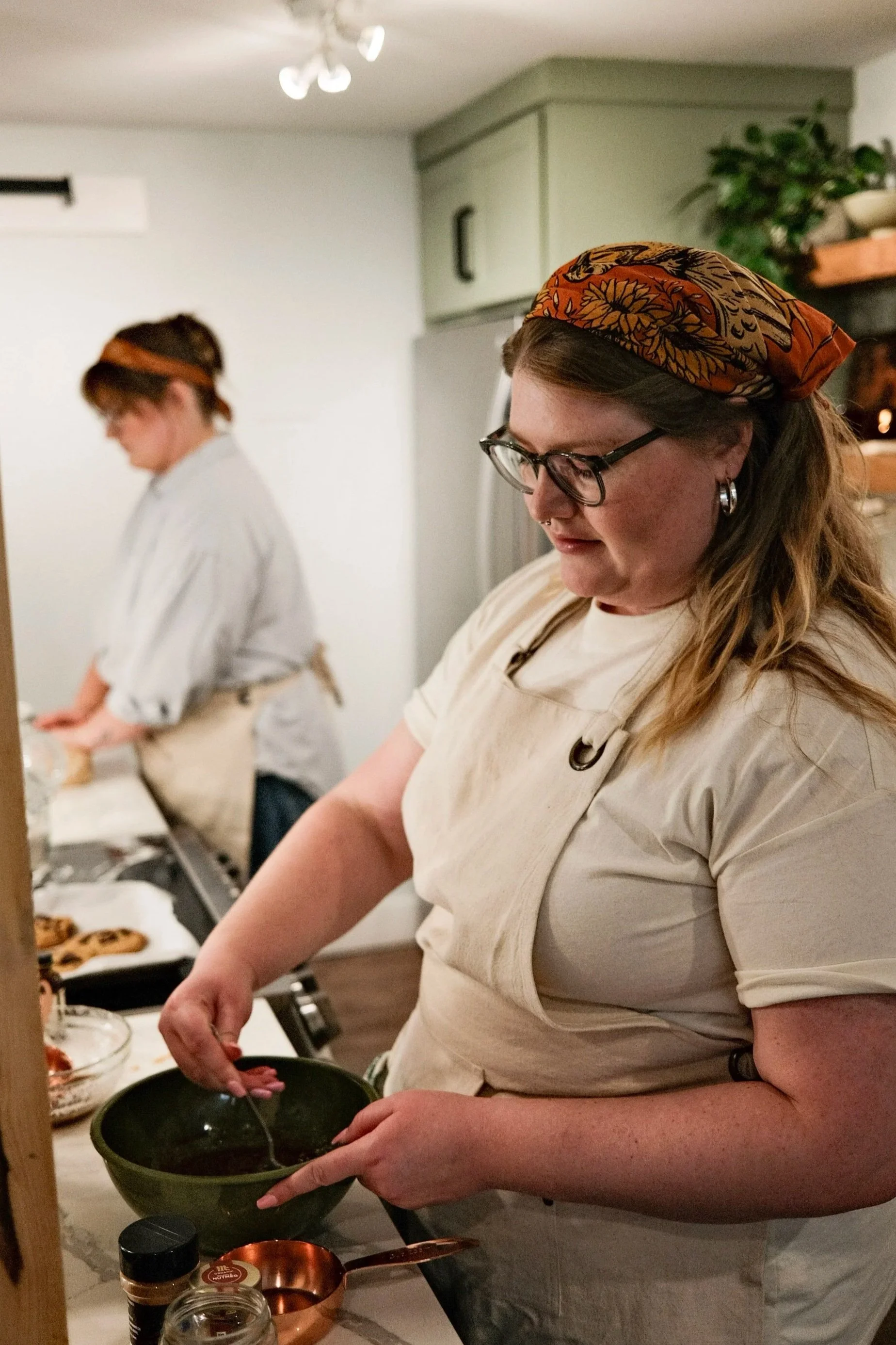 A woman wearing glasses, a headscarf, and a beige apron is preparing food in a kitchen. In the background, another person with a headscarf and apron is also working.