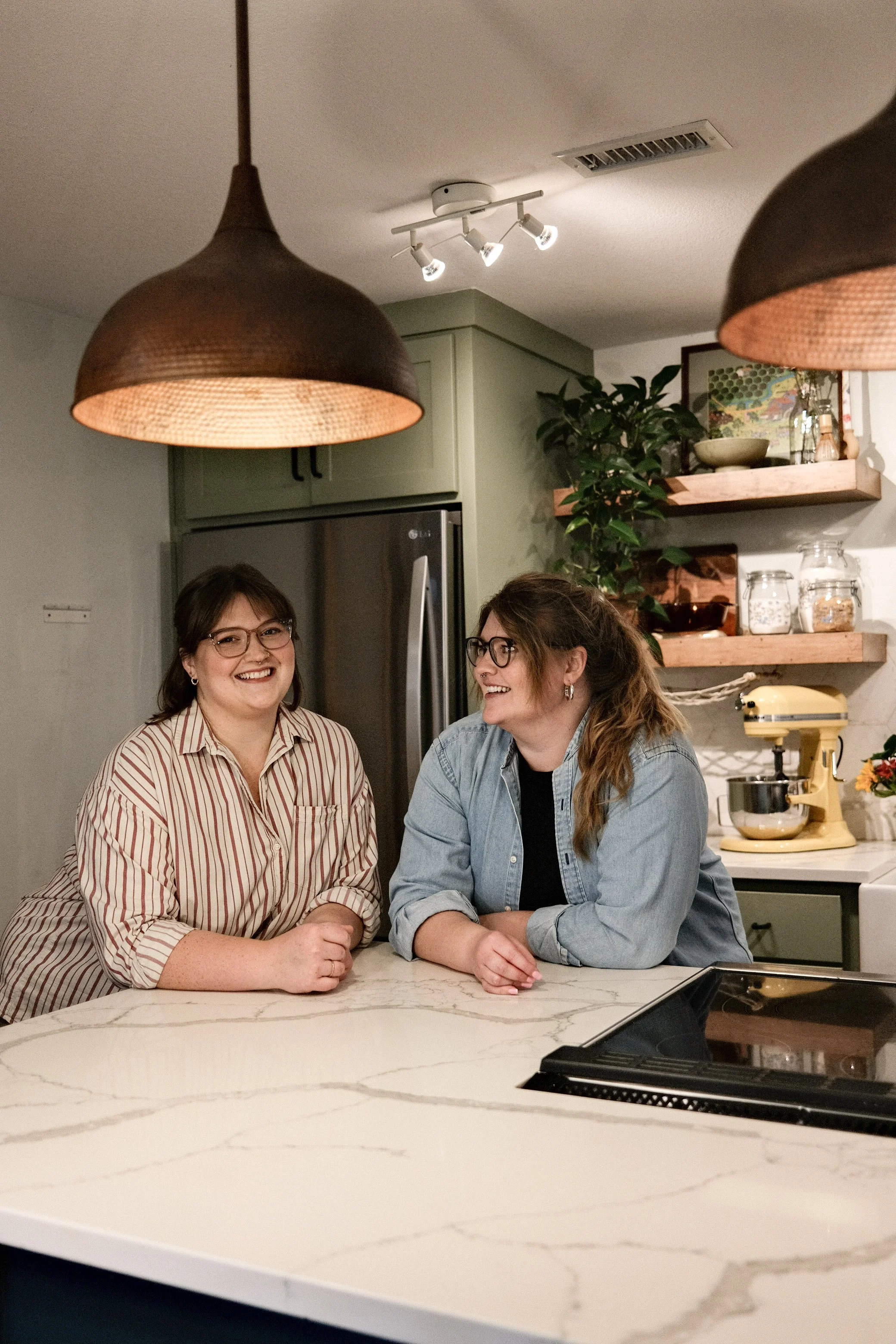 Two smiling women with glasses sitting at a marble kitchen island, with kitchen shelves, plants, and appliances in the background, lit by hanging and ceiling lights.
