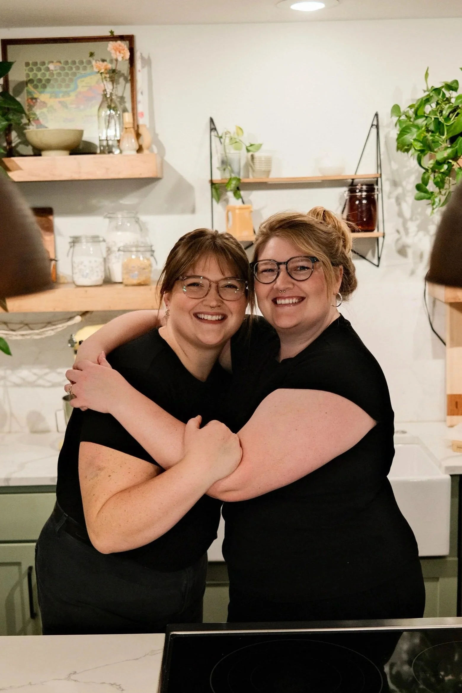 Two women in black shirts hugging and smiling in a kitchen with shelves and plants in the background.