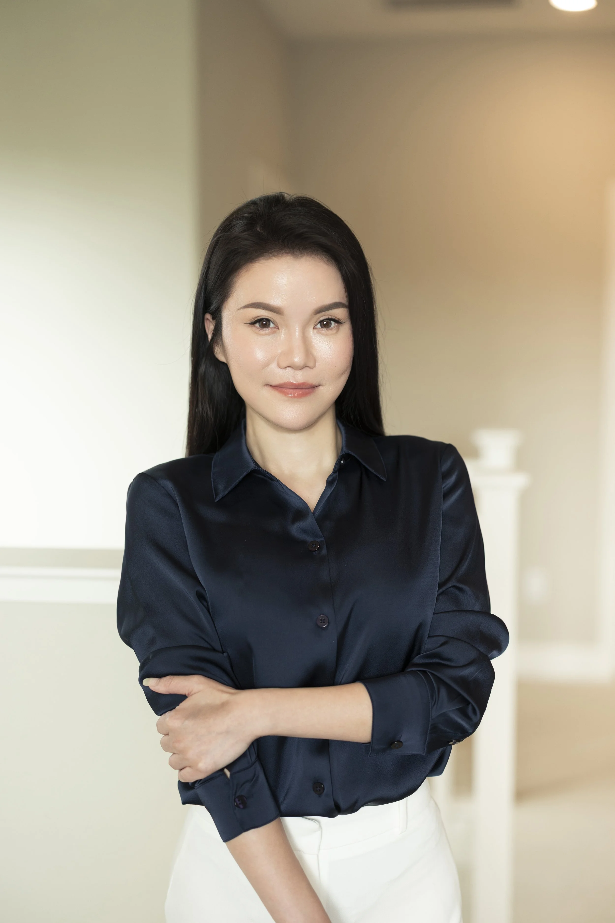 A young woman with straight, dark hair and light complexion standing indoors with her arms crossed, wearing a navy blue button-up shirt and white pants, smiling softly at the camera.