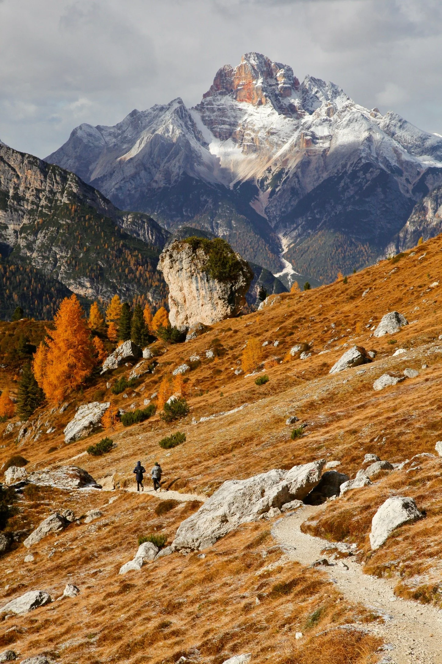 Deux personnes marchant sur un sentier dans un paysage de montagnes avec des arbres aux couleurs d'automne et un sommet enneigé en arrière-plan.