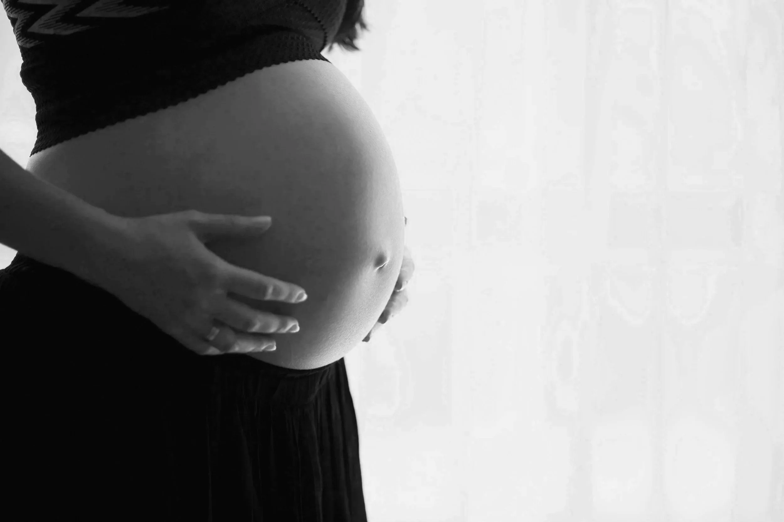 Black-and-white close-up of a pregnant woman cradling her belly by a bright window
