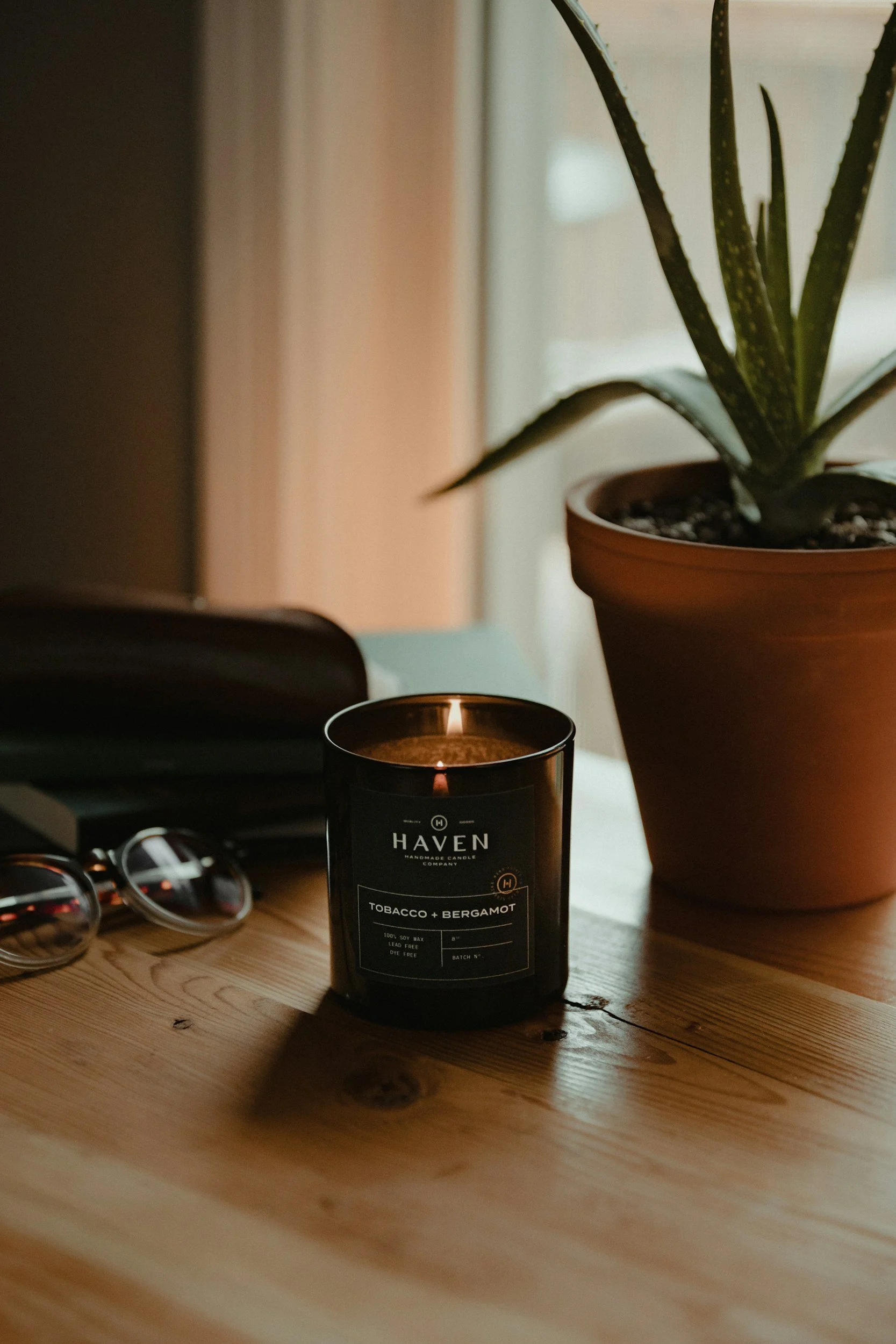 A lit black candle labeled ''Haven Handmade Candle Company, Tobacco + Bergamot'' on a wooden surface. Behind it, there's a potted aloe Vera plant on the right side, a pair of sunglasses on the left side, a dark object in the background, and part of a pair of scissors visible on the left.