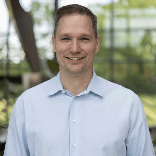 Smiling man in a light blue dress shirt in an indoor space with glass windows and green trees outside.