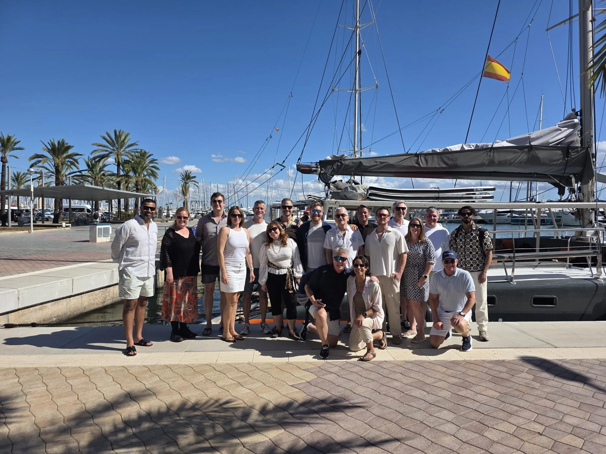 A group of CommodityFirst employees on a dock near sailboats and yachts with masts and a Spanish flag, palm trees, and a clear blue sky in the background.