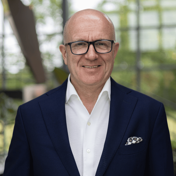 A bald man wearing glasses, a white shirt, and a navy blue suit jacket, smiling in an indoor setting with blurred green trees in the background.