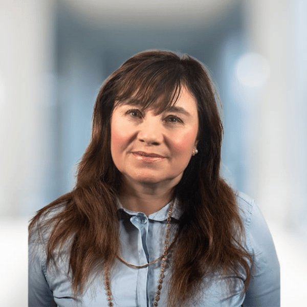 A woman with long brown hair and a blue collared shirt standing indoors, smiling softly at the camera.