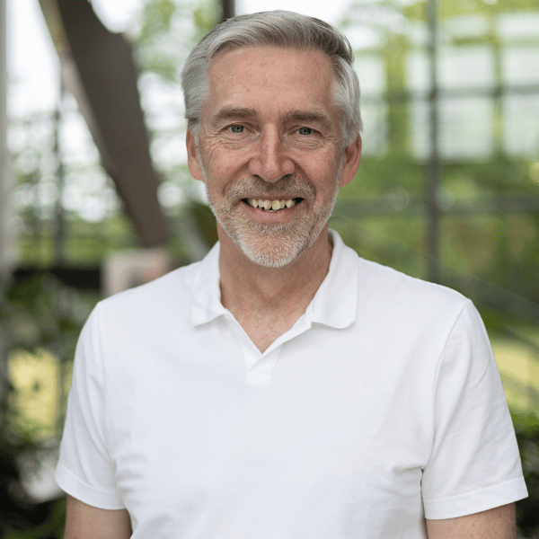 A smiling older man with gray hair and a beard wearing a white polo shirt. Background features a modern indoor setting with large windows and greenery outside.