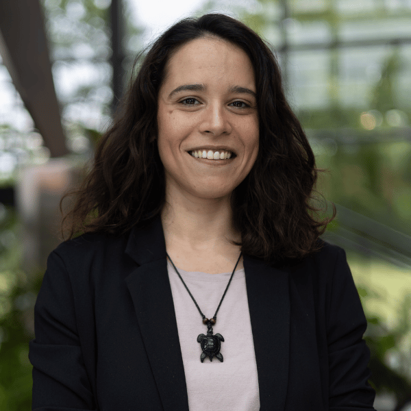 A woman with long dark curly hair smiling, wearing a black blazer and a light-colored shirt with a black turtle-shaped necklace, standing outdoors with a blurred background of greenery and glass structures.