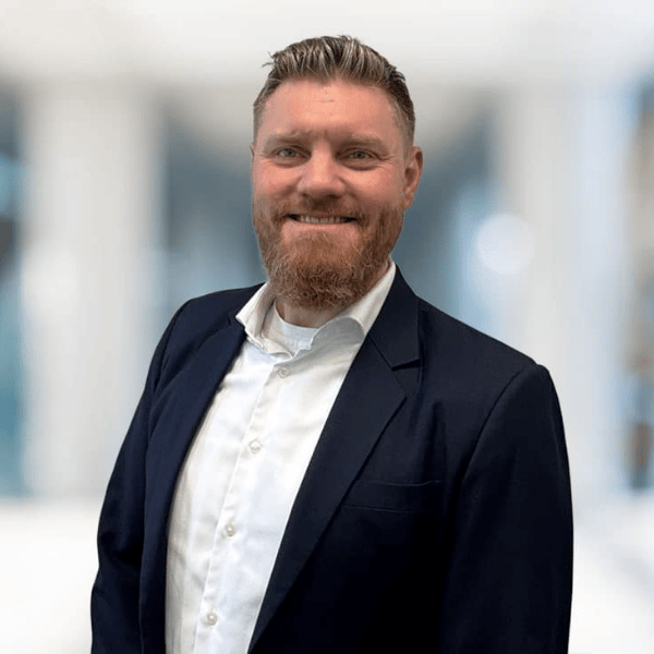 A man with a beard and short hair, wearing a dark suit jacket and white shirt, standing in a bright, blurred indoor setting, smiling at the camera.