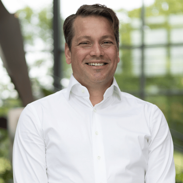 Portrait of a smiling man wearing a white button-up shirt, standing indoors with large windows and greenery outside in the background.