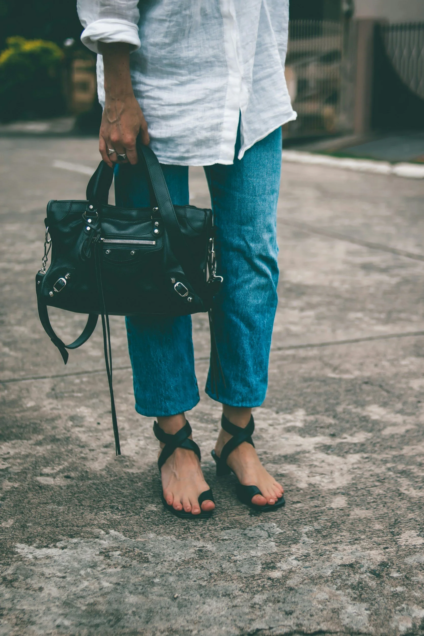 Person standing barefoot on a paved surface, holding a black handbag, wearing a white shirt and blue jeans.