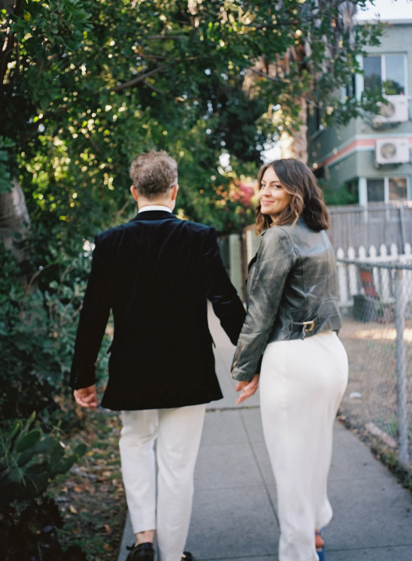 A man and woman walking away from the camera; green trees and buildings are in the background. The woman turns her head around to look at the camera.
