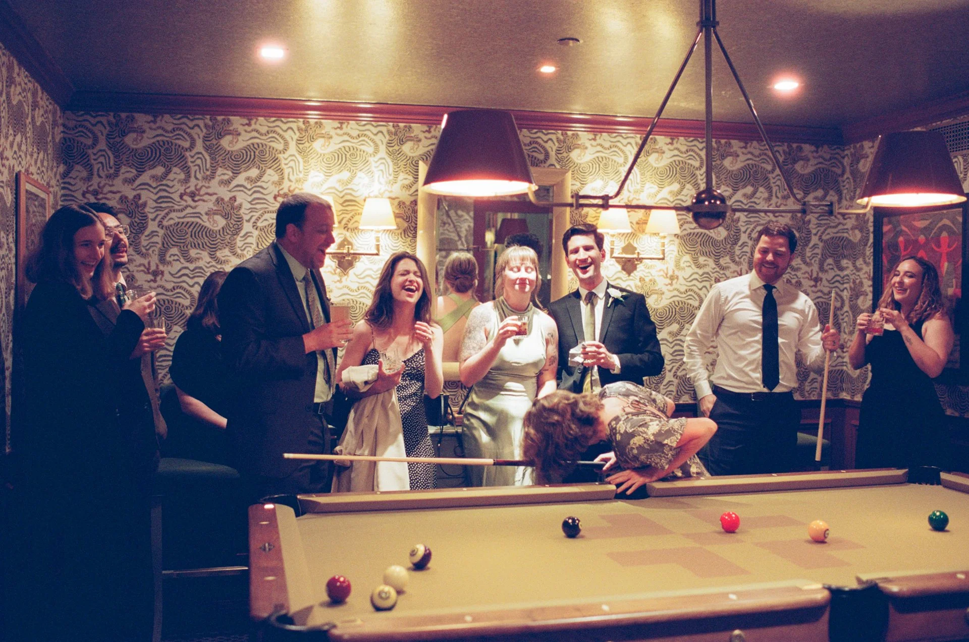 Group of people in formal attire enjoying a social gathering around a pool table in a decorated room. Several individuals are holding drinks and laughing.
