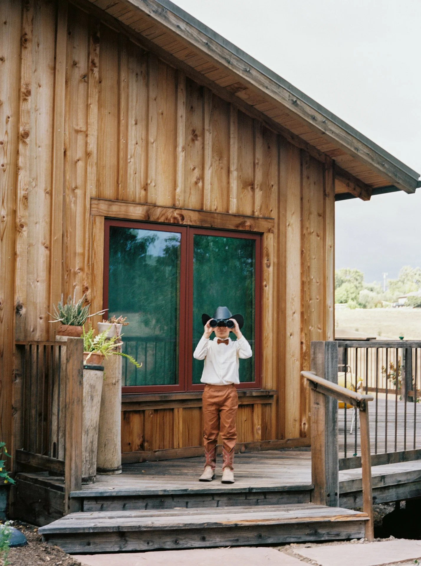 A young boy stands on a wooden deck holding binoculars to his eyes, wearing a cowboy hat, a white shirt, an orange bow tie, brown pants, and cowboy boots, against the backdrop of a wooden building and cloudy sky.