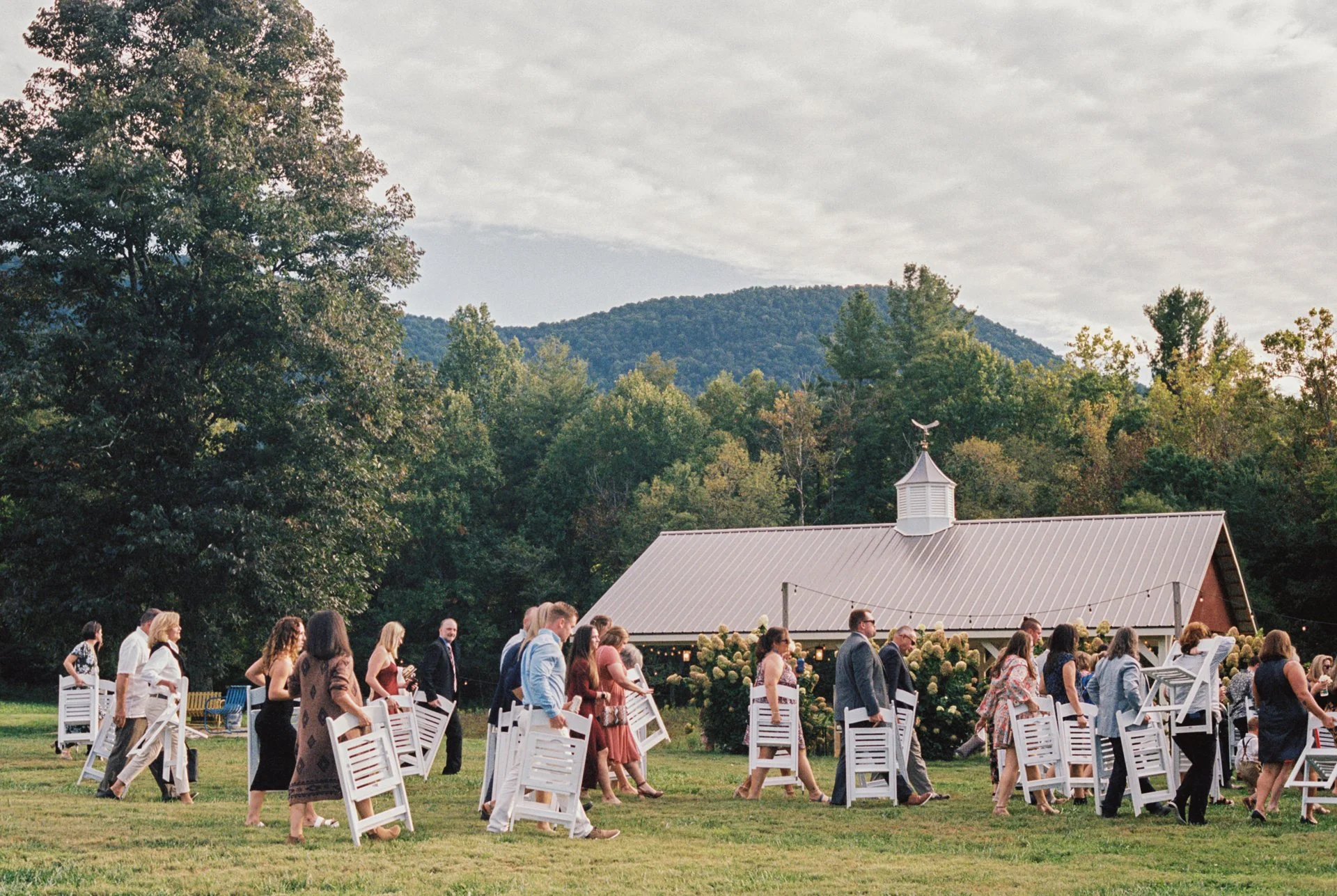 People attending an outdoor wedding ceremony on a grassy field with a rustic barn with a weather vane, trees, and a mountain in the background under cloudy sky. They are carrying white folding chairs.