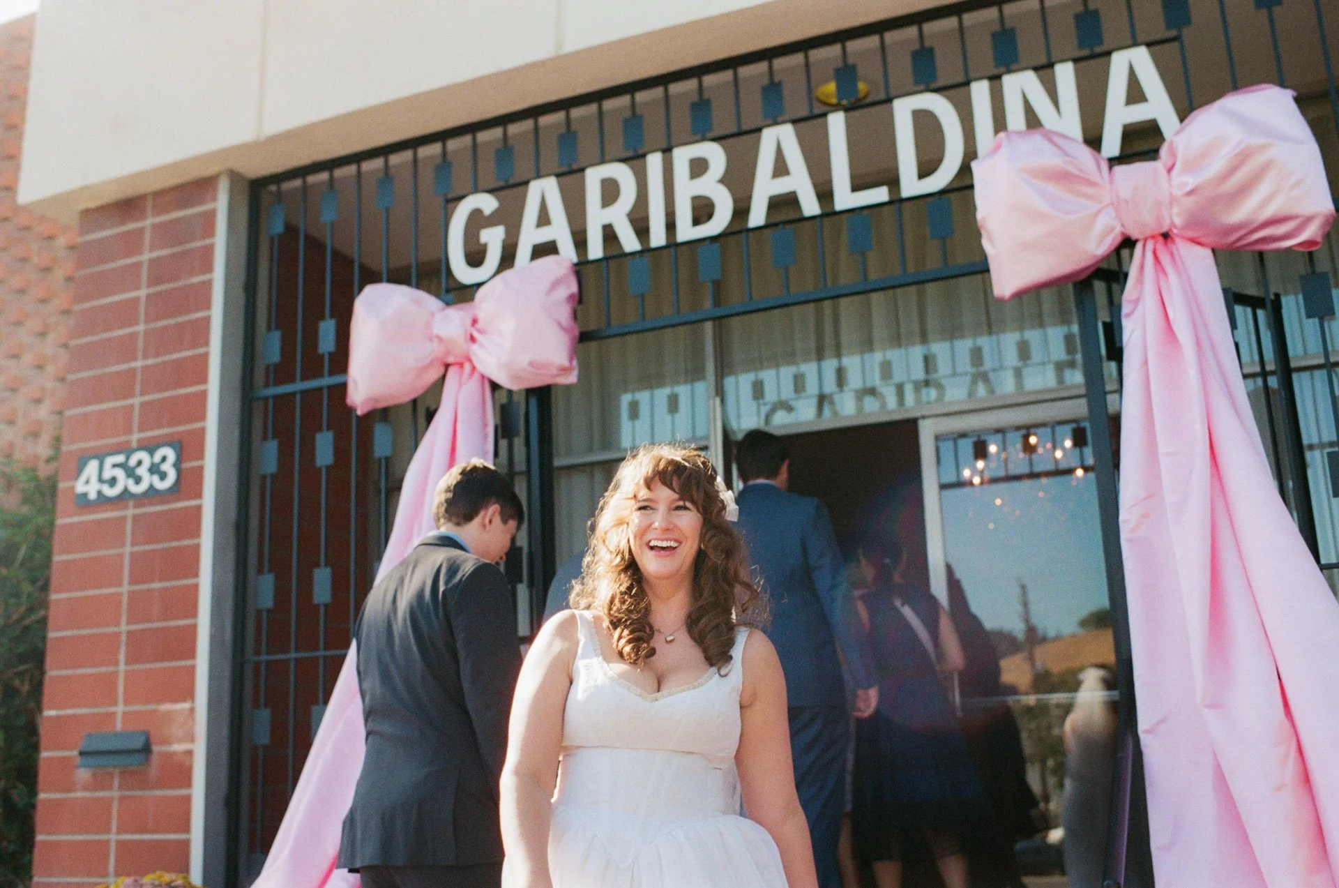A woman in a white wedding dress smiling in front of a decorated wedding venue with pink bows on the gate, and people dressed in formal attire in the background.