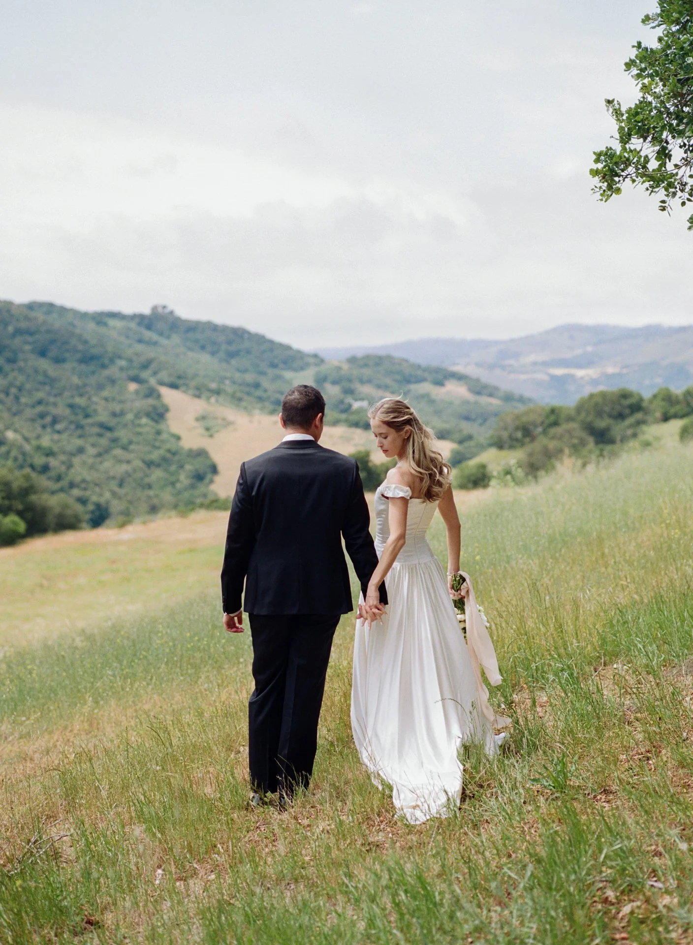 A bride and groom holding hands in a green field with rolling hills in the background.