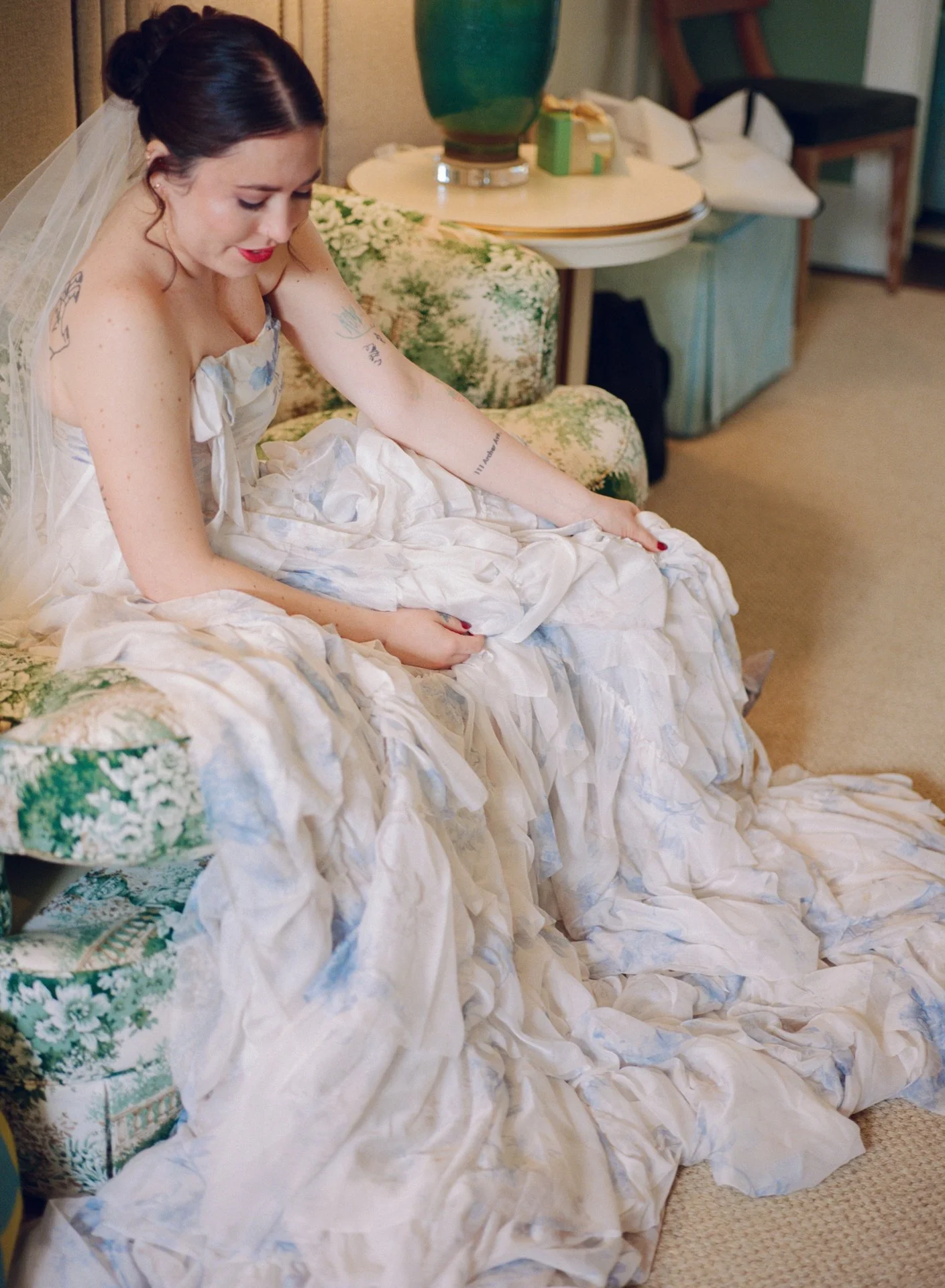 A woman dressed as a bride sitting on a couch, looking down with a gentle smile, as she fixes her white and blue wedding dress. 