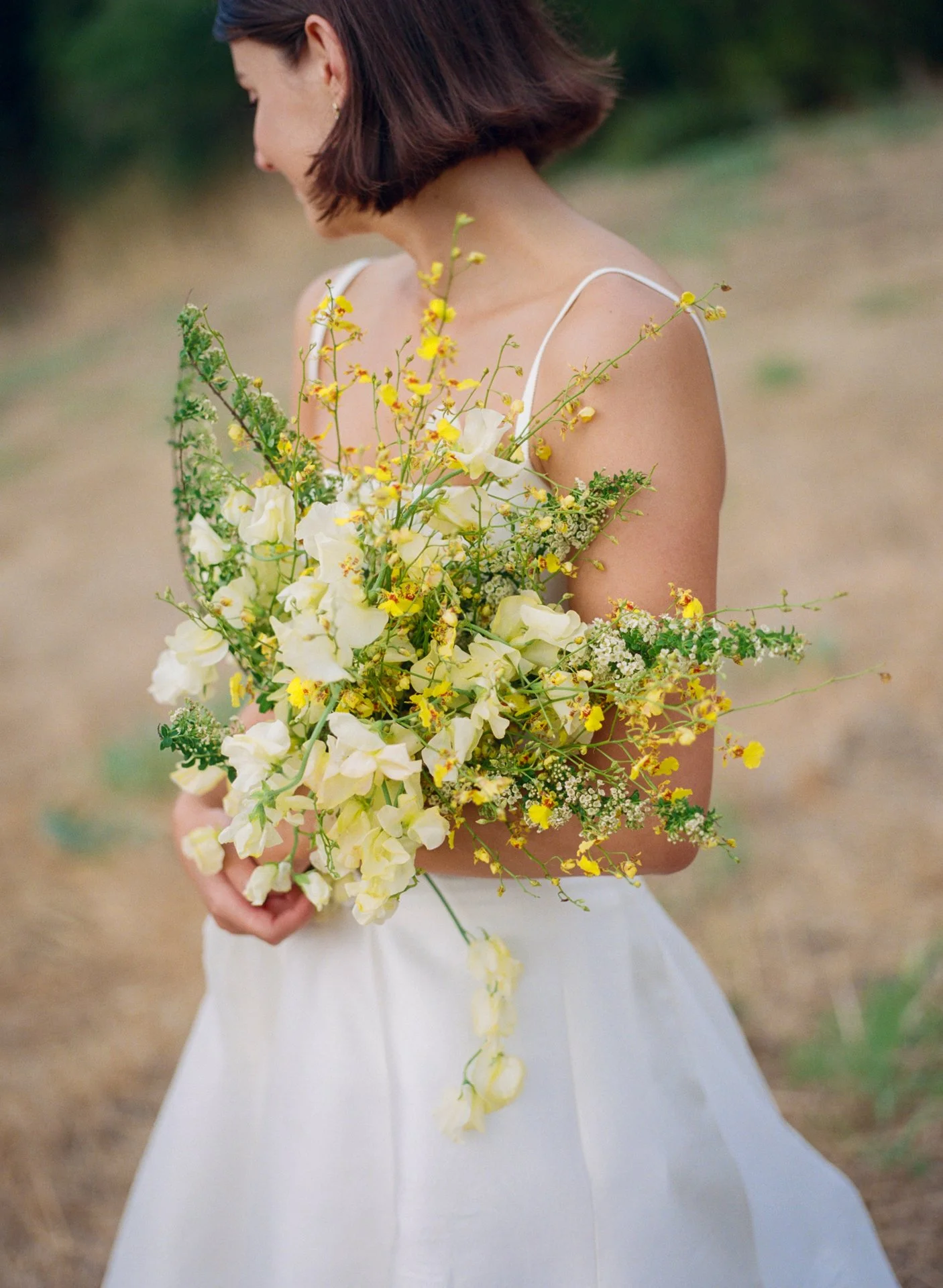 A woman wearing a white wedding dress holding a large bouquet of yellow and cream flowers, in a field.