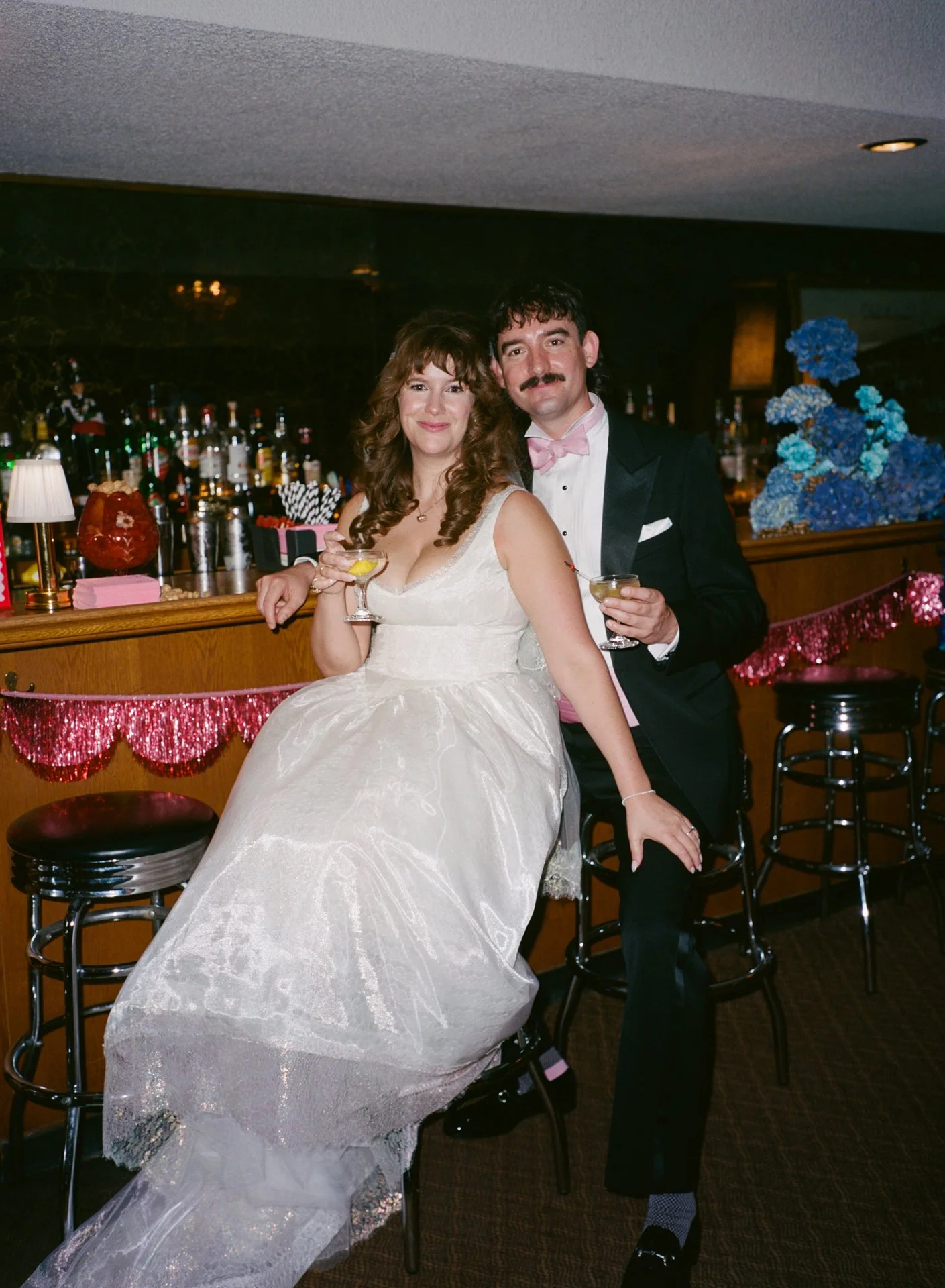 A bride and groom at a bar during their wedding celebration, holding drinks and smiling at the camera.