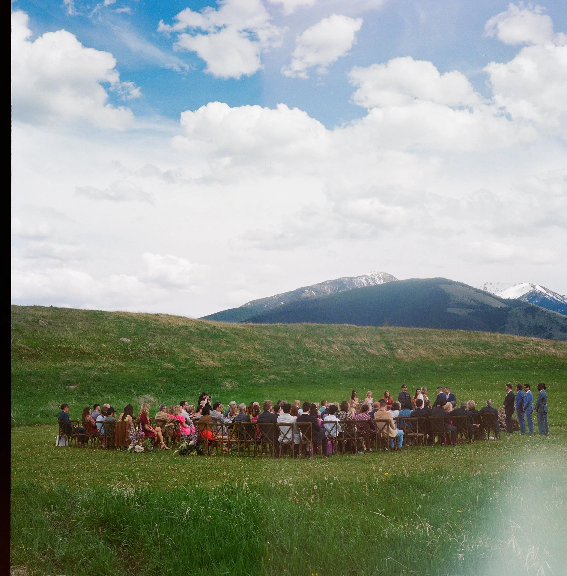 People attending an outdoor wedding ceremony on a hillside in front of a mountain range under a cloudy sky.