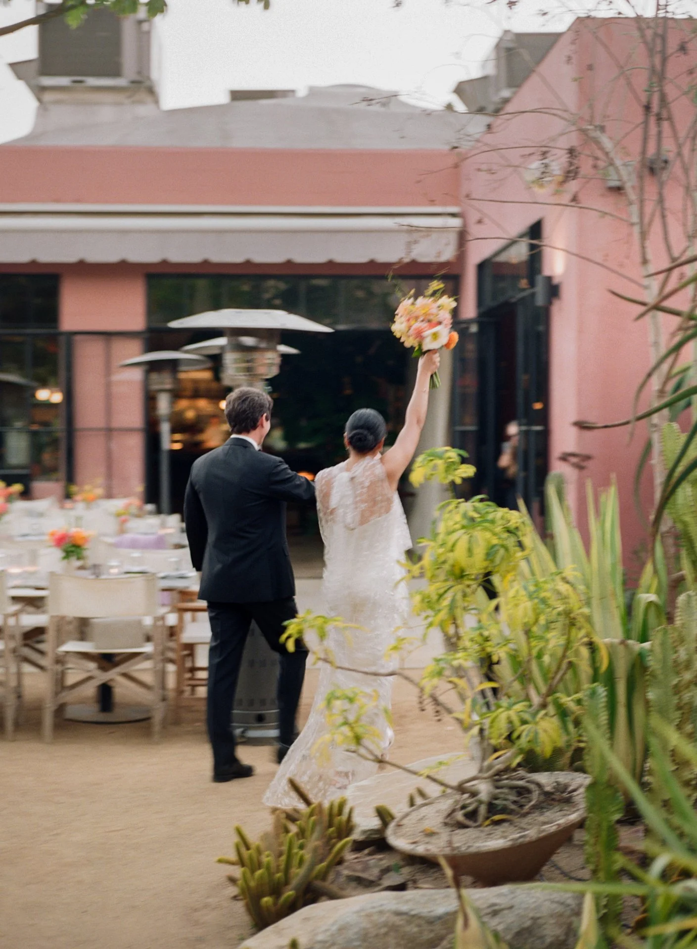 A bride holding a bouquet of flowers in the air, walking with a groom dressed in a suit in an outdoor courtyard or garden, surrounded by plants and tables, with a pink building in the background.