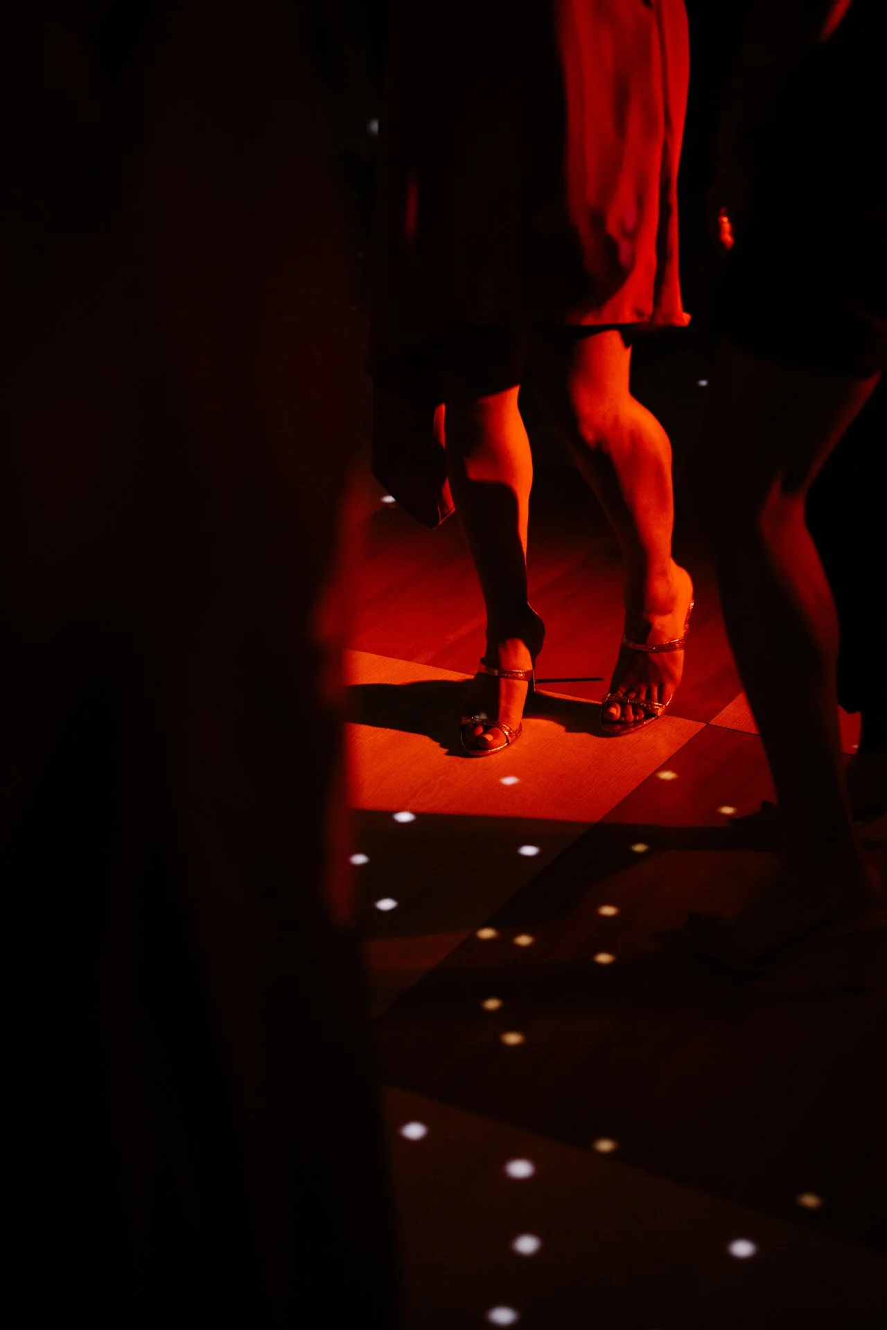 Close-up of a woman's legs in high heels on a dance floor with red lighting and disco ball casting reflections onto the floor.