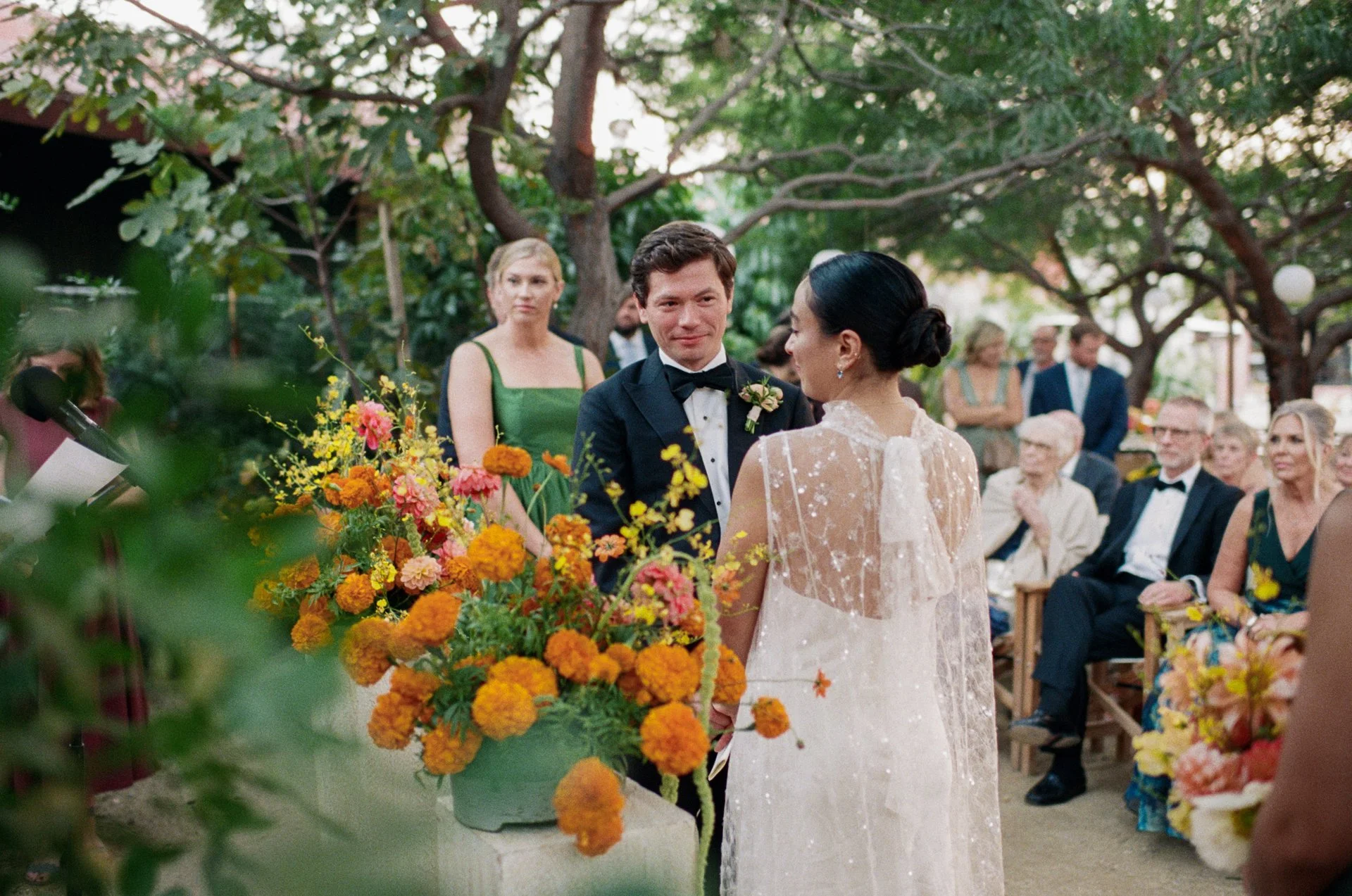 A wedding ceremony outdoors surrounded by trees, with a bride and groom exchanging vows in front of seated guests. The bride wears a lace wedding dress, and the groom is in a black tuxedo. A floral arrangement with orange and yellow flowers decorates