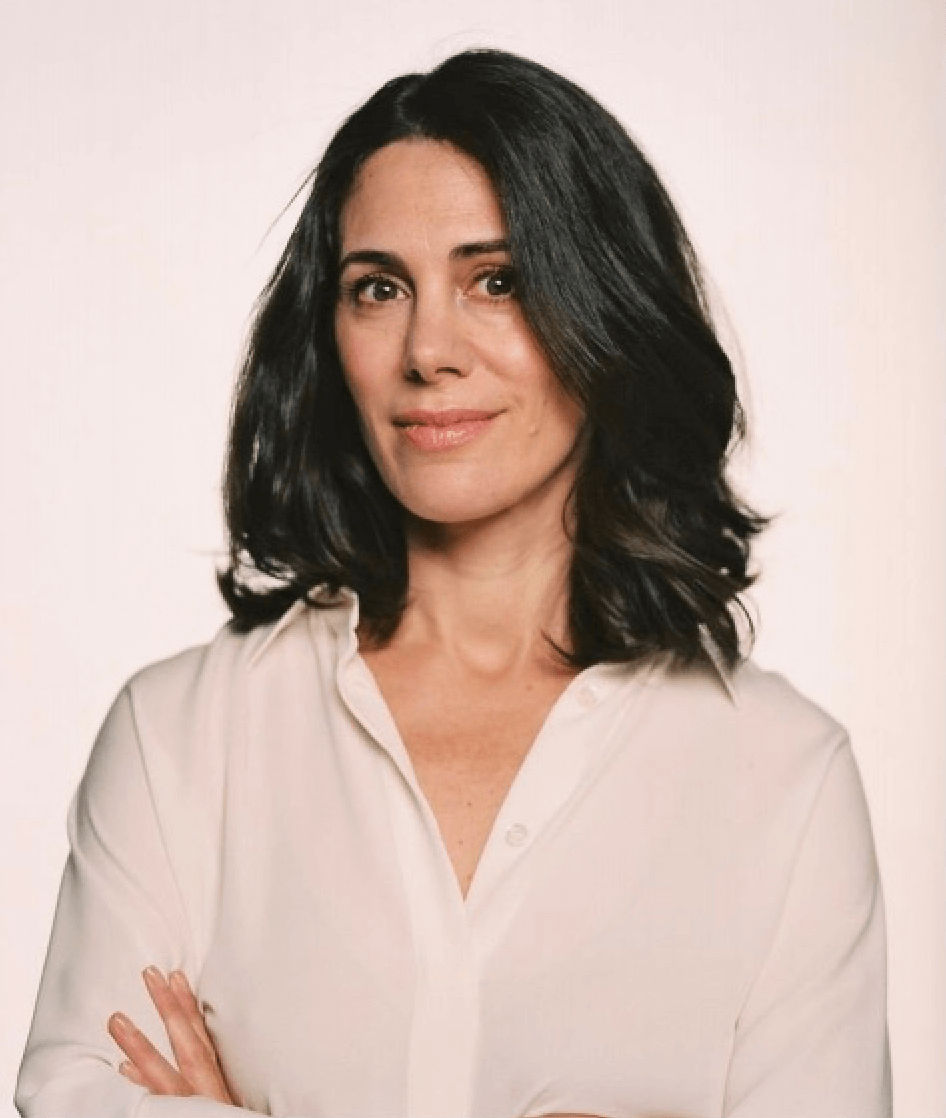A woman with shoulder-length black hair wearing a cream-colored blouse, standing against a light background with her arms crossed, smiling softly.