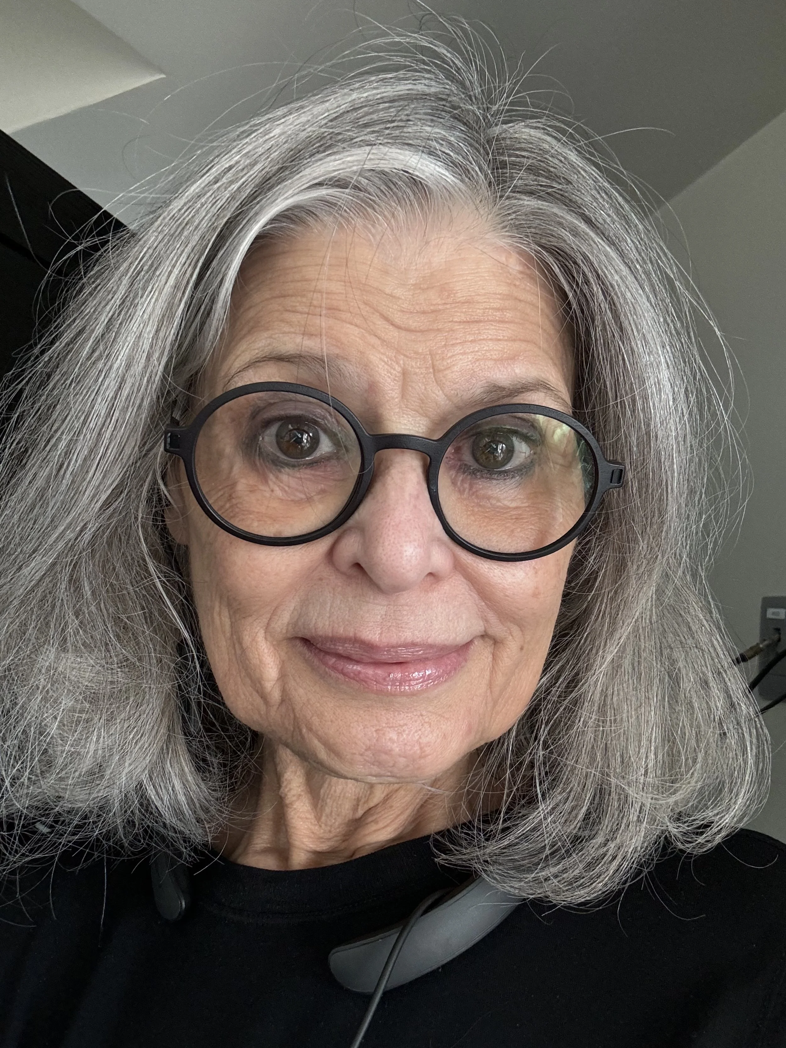 Close-up of a smiling woman with gray hair, wearing black glasses, sitting at a desk with a phone headset, in an indoor setting.