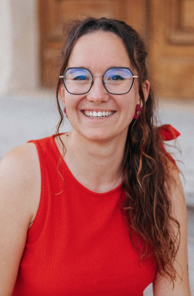 Une femme souriante portant des lunettes et un haut rouge, avec des cheveux longs et ondulés, assise à l'extérieur.