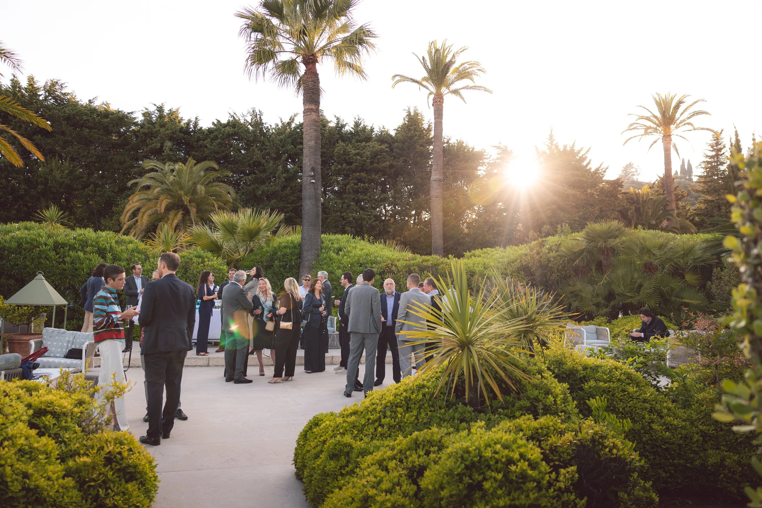 Groupe de personnes en costume discutant dans un jardin avec palmiers et soleil couchant.