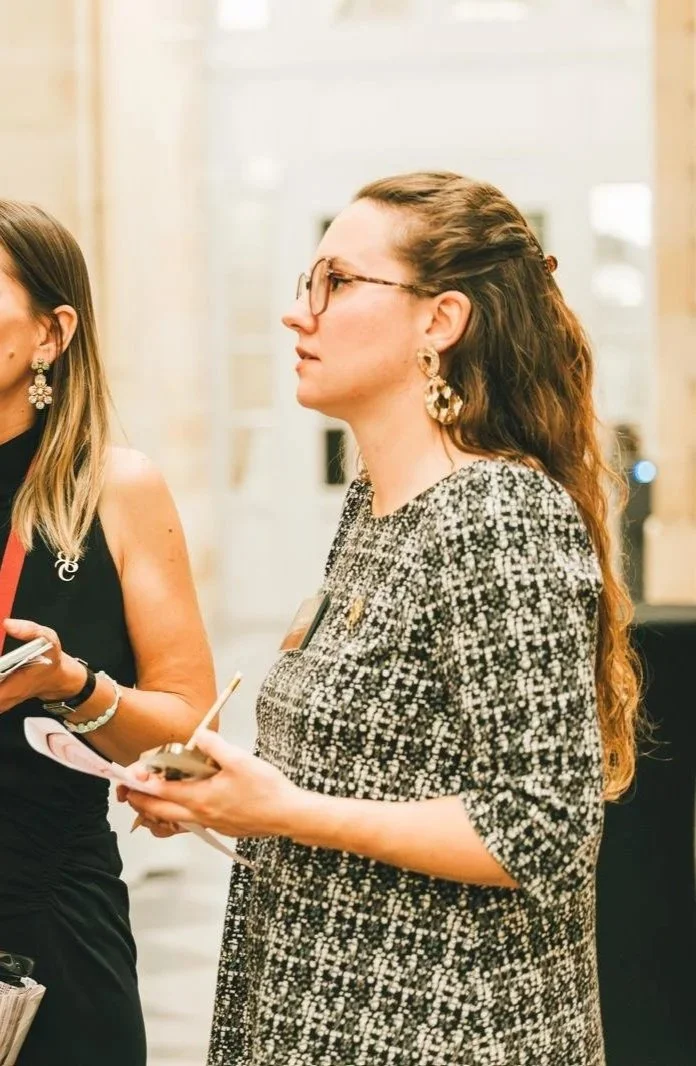 Femme portant des lunettes, une robe à motifs noirs et blancs, avec de grosses boucles d'oreilles, tenant un carnet et un stylo, lors d'un événement intérieur.