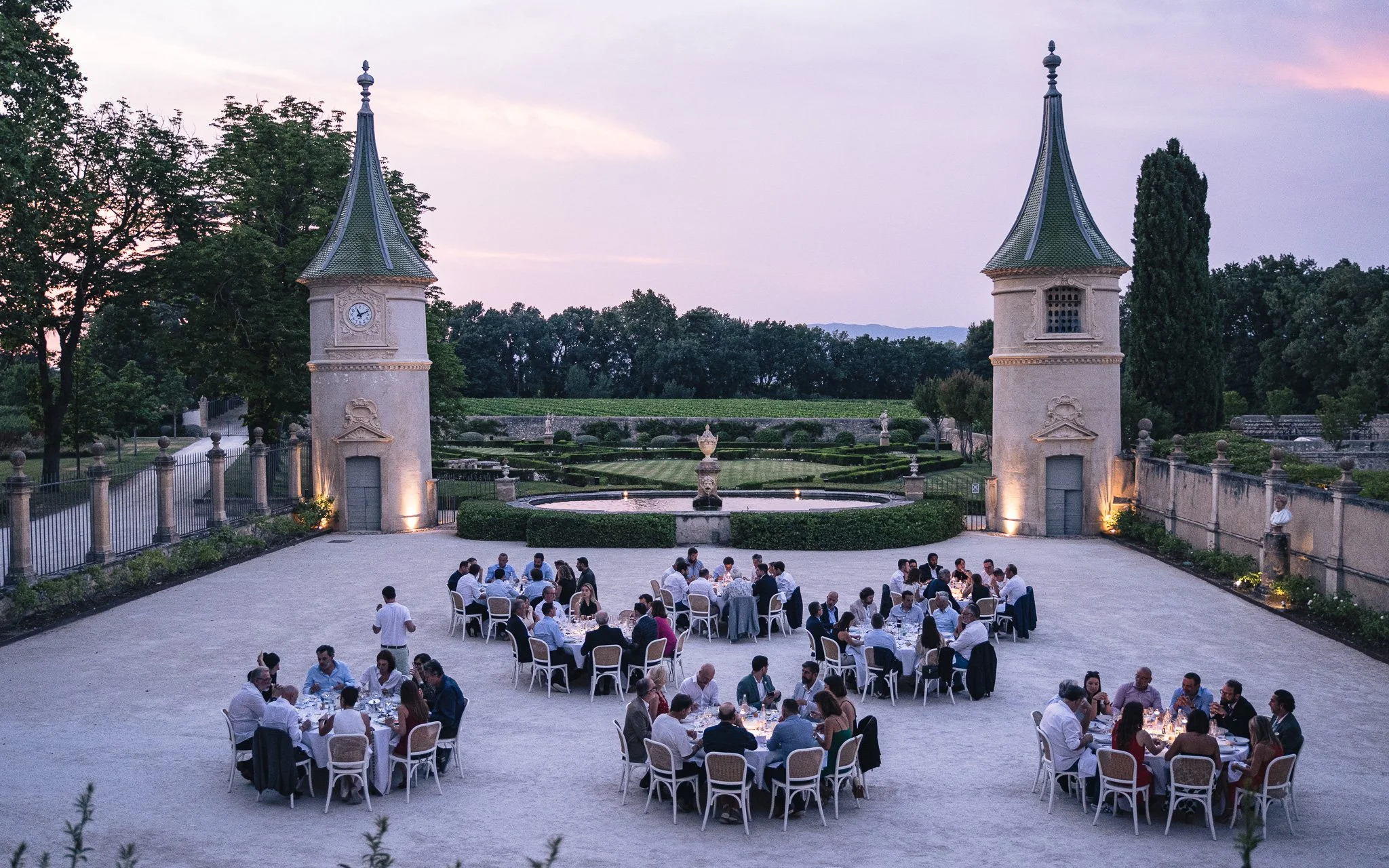 Dîner en plein air avec plusieurs tables et invités dans un jardin, avec deux tours de château en arrière-plan, le tout lors du crépuscule.