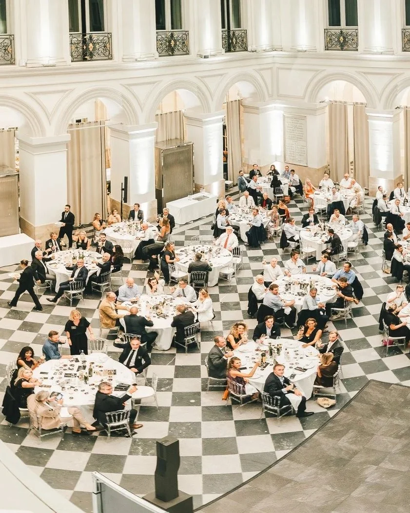 Banquet dans une grande salle élégante avec vieil architecture, tables rondes avec invités en tenue formelle, sol en carreaux noirs et blancs.