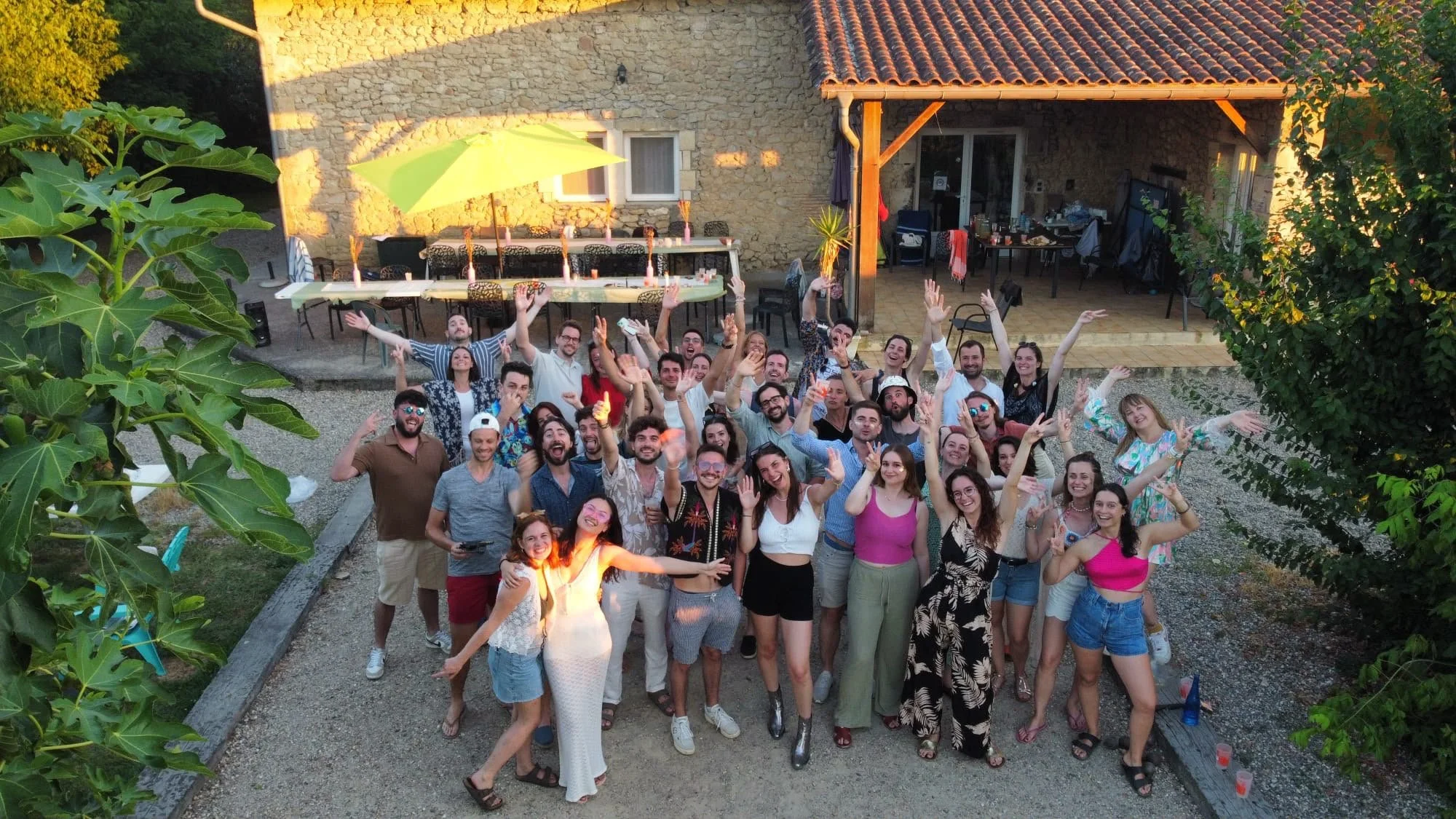 groupe de personnes souriantes et joyeuses lors d'une fête en extérieur dans un jardin, avec une table décorée et une villa en pierre en arrière-plan