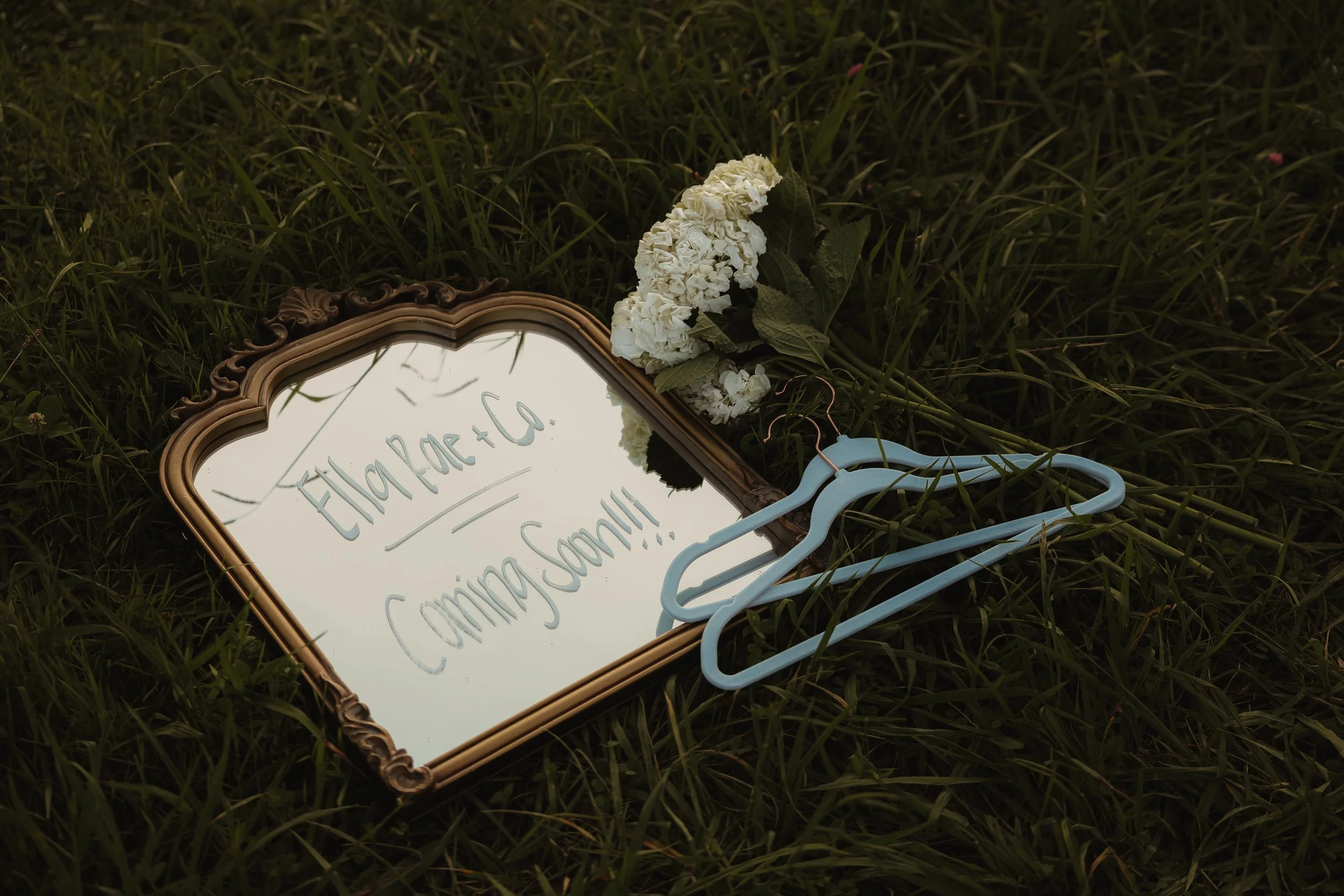 A mirror with a decorative gold frame lying on the grass, reflecting handwritten text that says 'Elliot, small! Come!!' with a bouquet of white flowers resting on the mirror's edge and a light blue hanger nearby.
