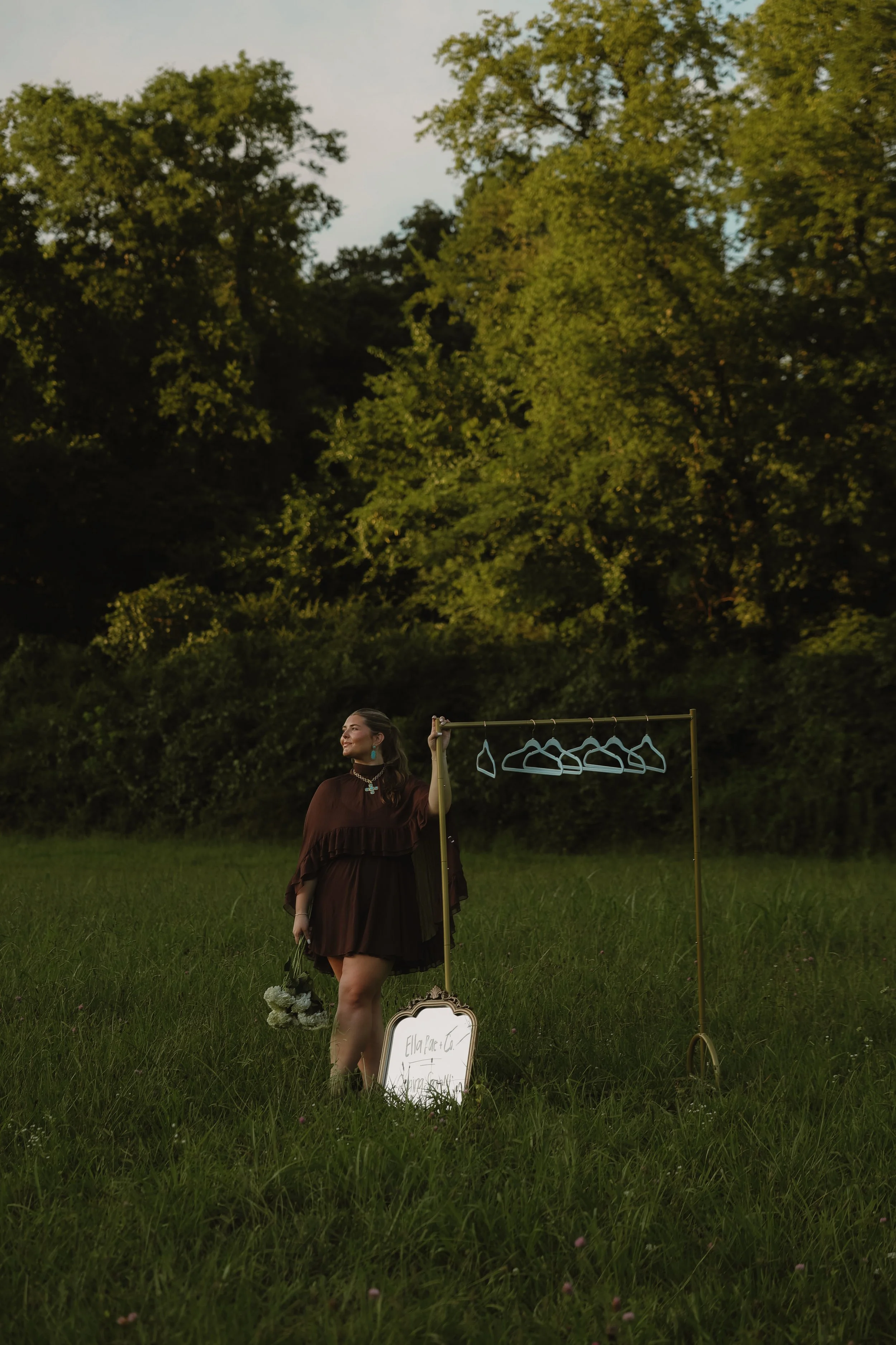 A woman dressed in a dark brown dress standing in a grassy field holding a clothing rack with clothes hangers. There is a sign at her feet and a bouquet of white flowers. The background has green trees and foliage, and the sky is cloudy.