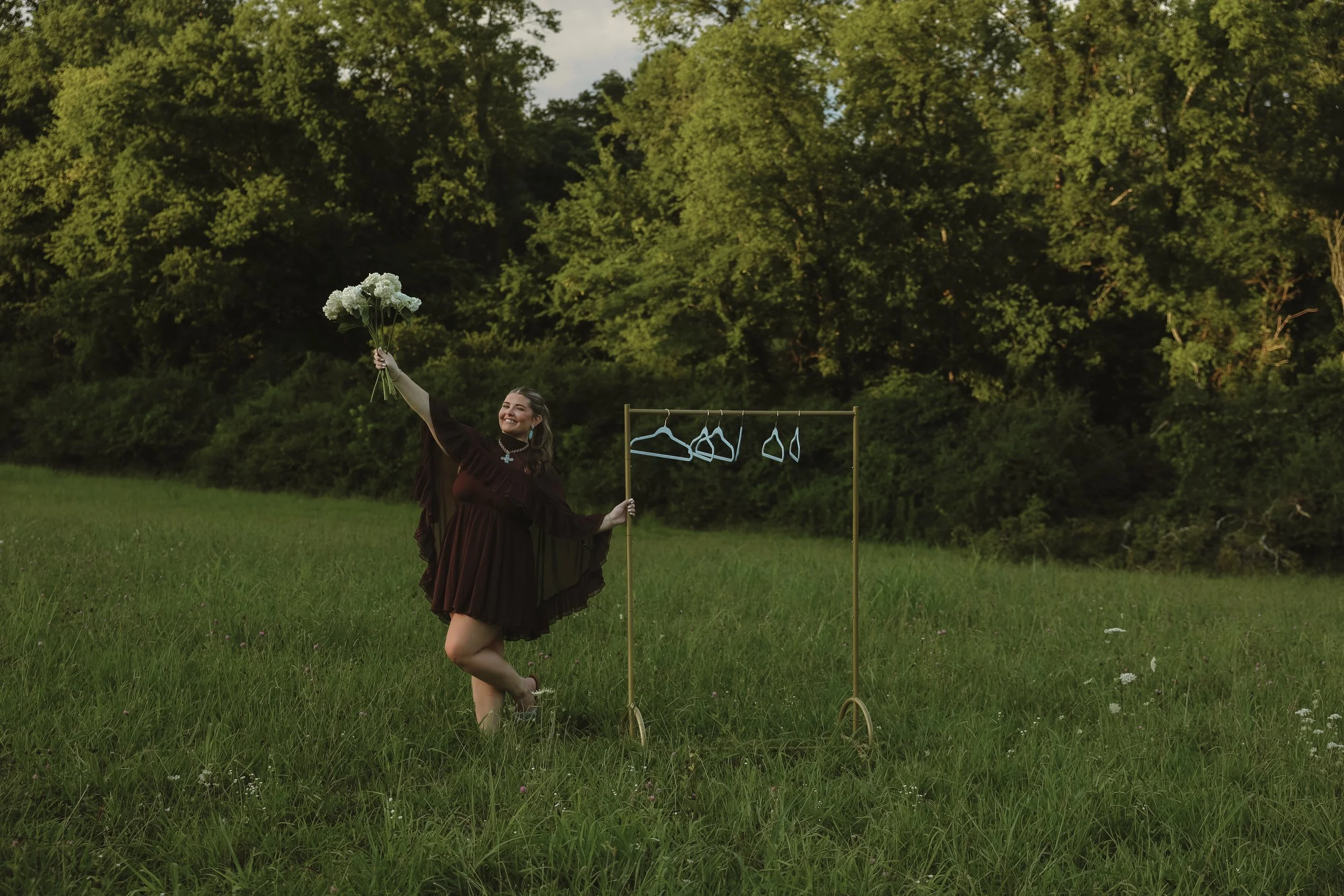 A woman in a brown dress dancing outdoors on a grassy field, holding a bouquet of white flowers in one hand, with a small clothing rack and hangers behind her, set against a background of green trees under a partly cloudy sky.