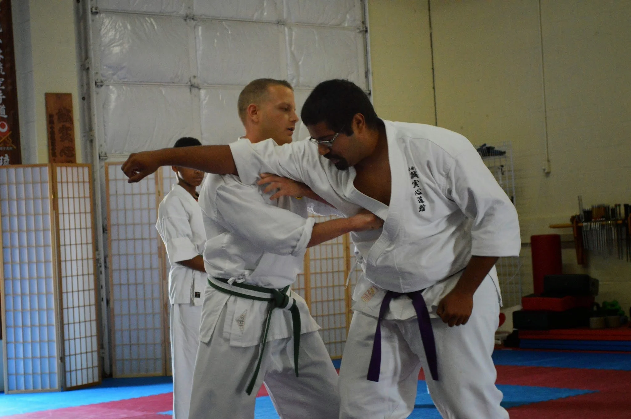 Two martial artists practicing judo or Brazilian Jiu-Jitsu in a dojo, both wearing white gis with colored belts, one with a green belt and the other with a purple belt, engaged in a training drill.