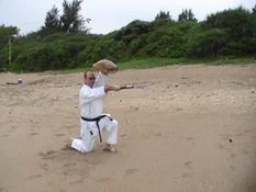 A person dressed in a white martial arts uniform kneeling on the sand beach, holding a large umbrella.