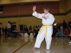 A person dressed in a white karate gi with a yellow belt performing a martial arts move in a gymnasium with other people in the background.