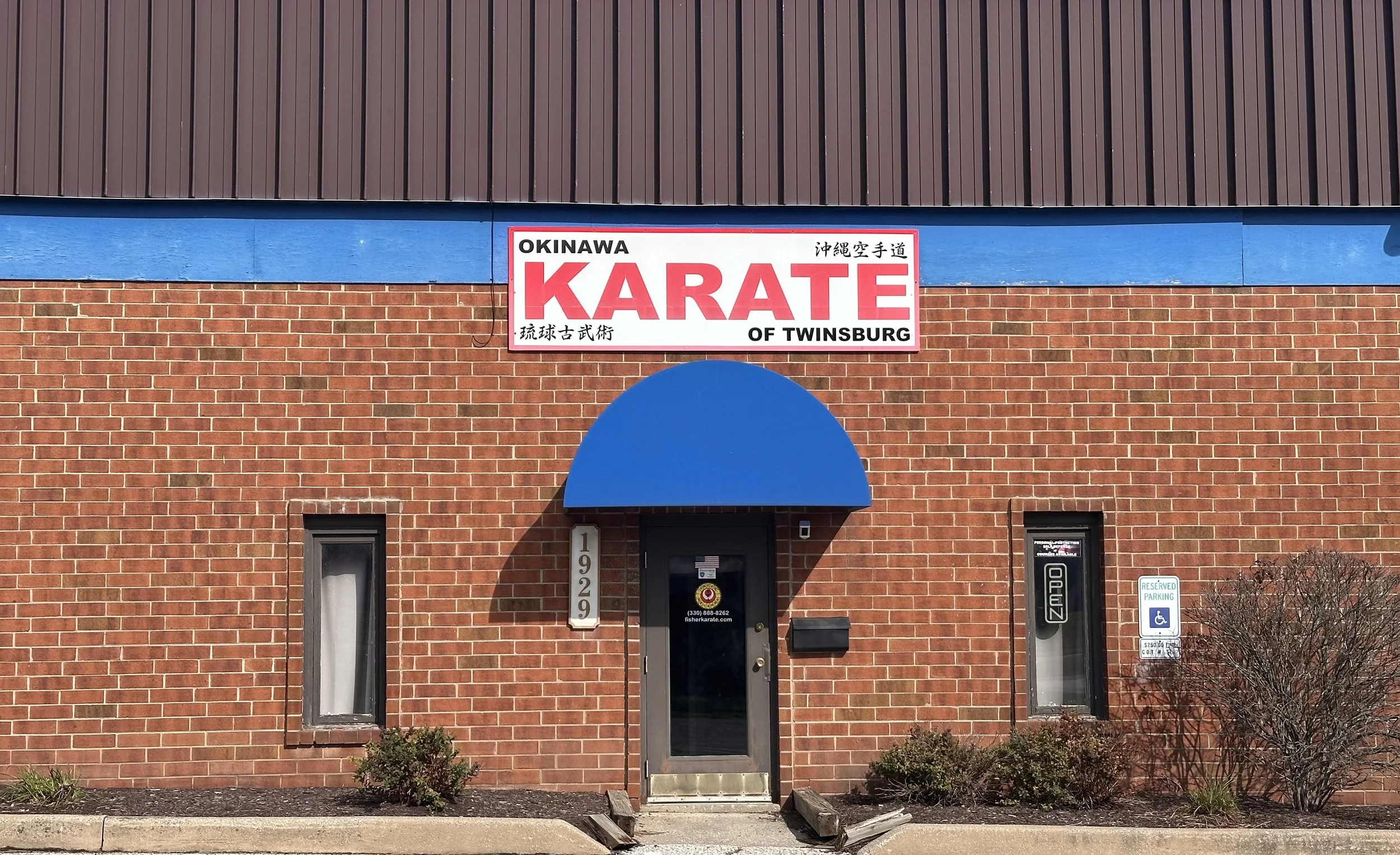 The exterior of a building with a brick facade and a large sign that reads 'Okinawa Karate of Twinsburg' in red and black letters, featuring Japanese characters, and a blue awning over the entrance.