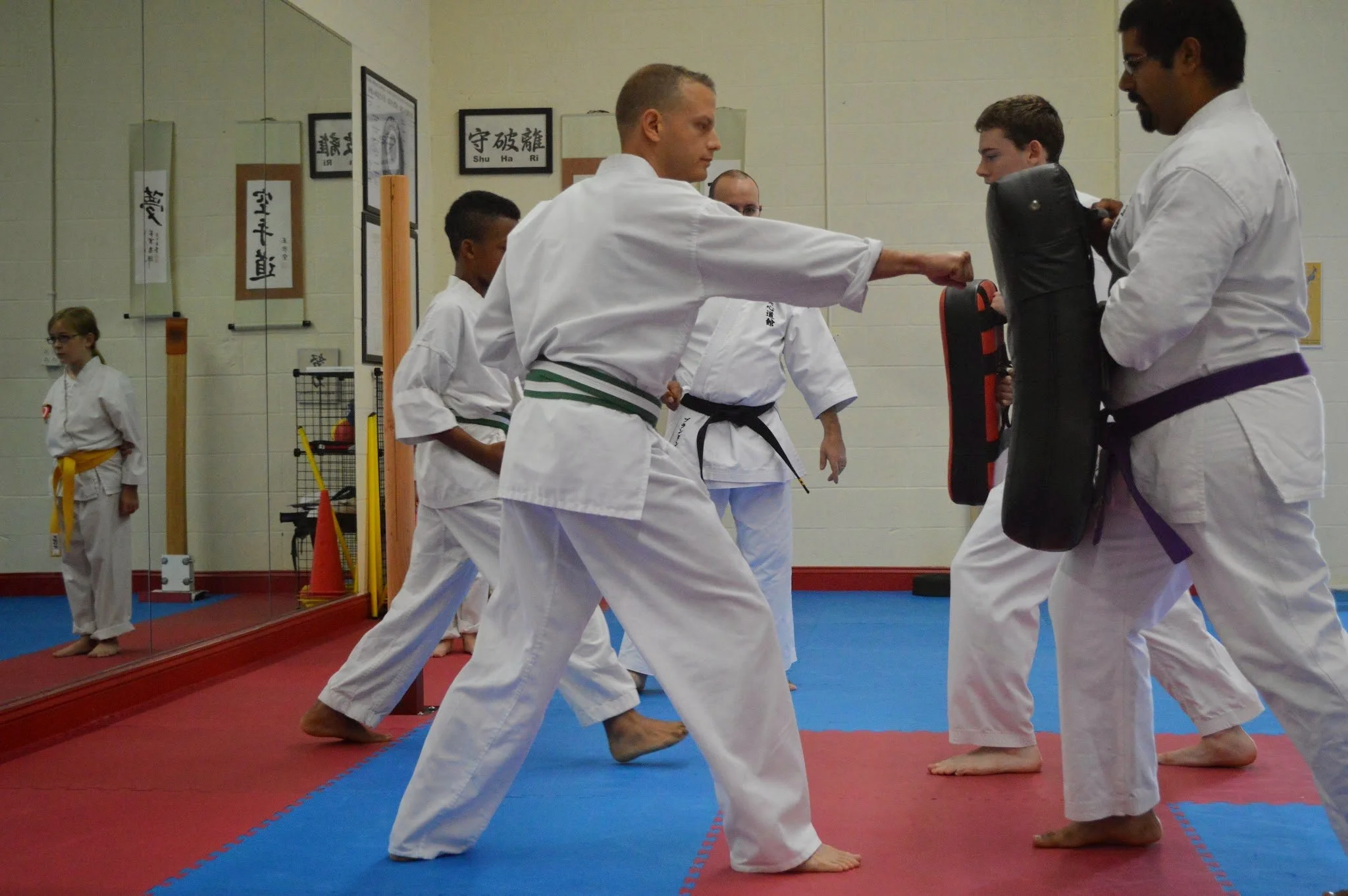 Martial arts students practicing techniques in a dojo, wearing traditional white uniforms and colored belts, with a mirror reflecting their activities.