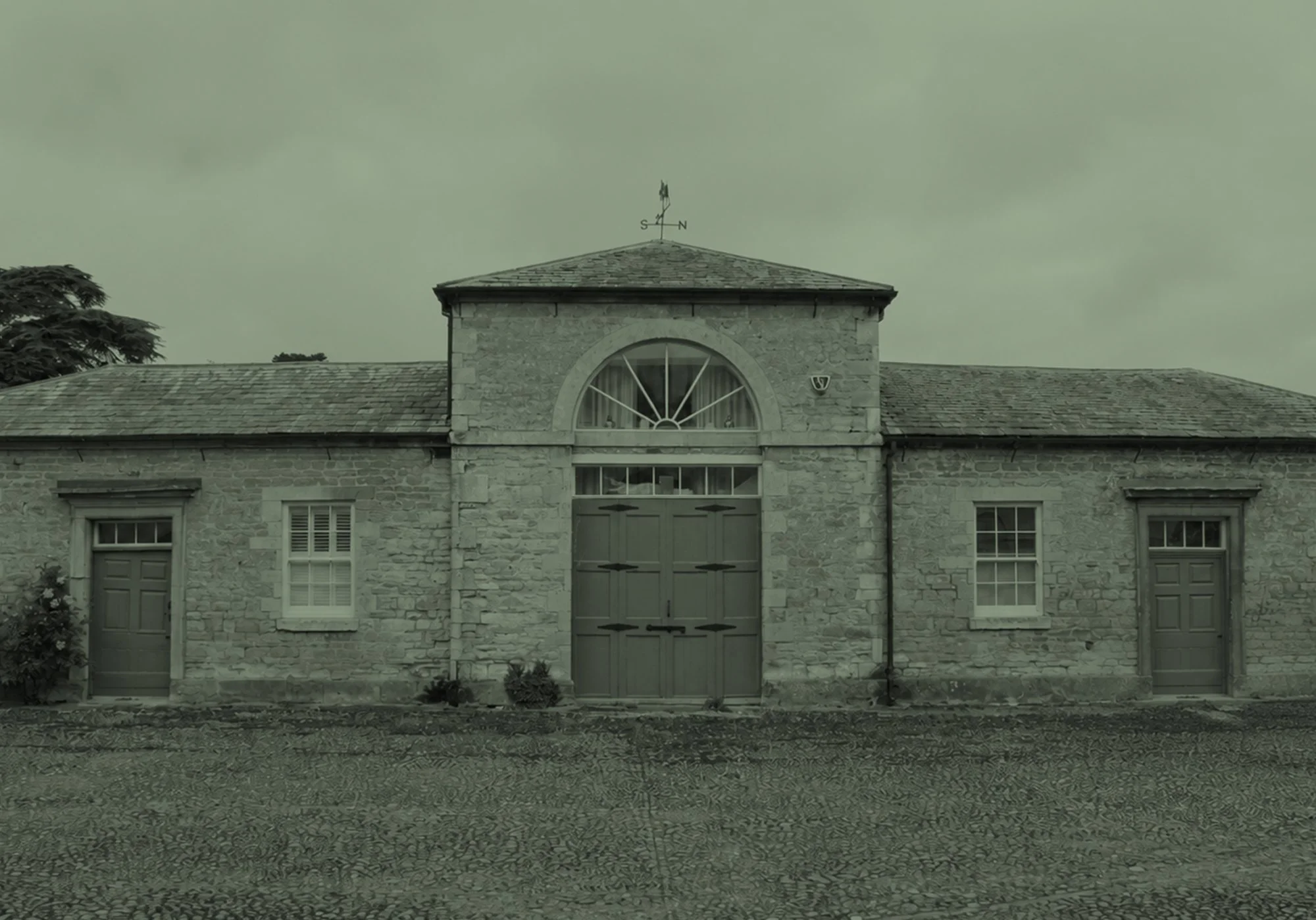 A stone building with three windows, a large arched window at the top, a door beneath it, and two smaller windows on either side, with a cobblestone driveway in front and trees in the background.
