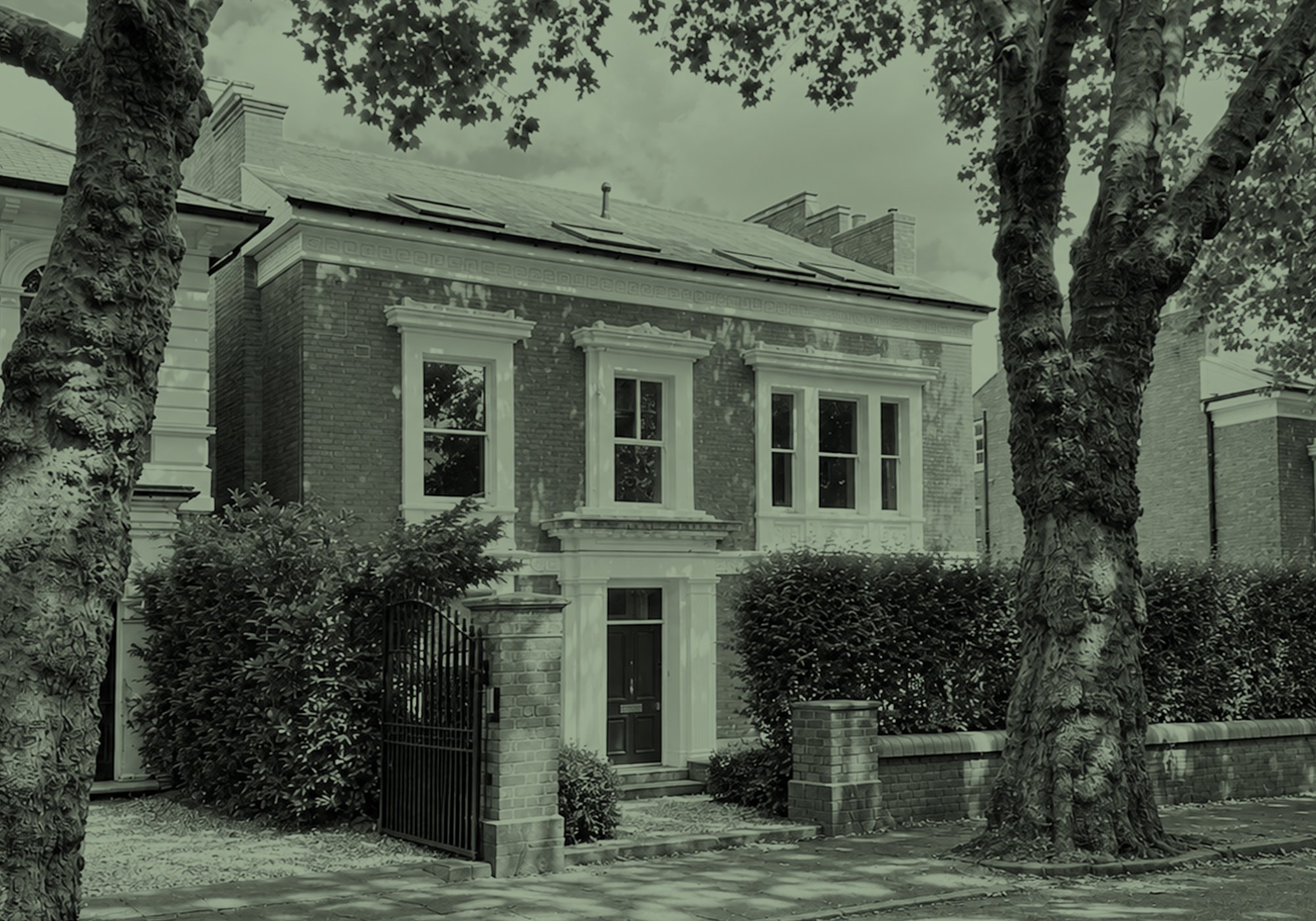A black and white photo of a two-story brick house with decorative window frames, a gated front yard, and large trees in front of the house.