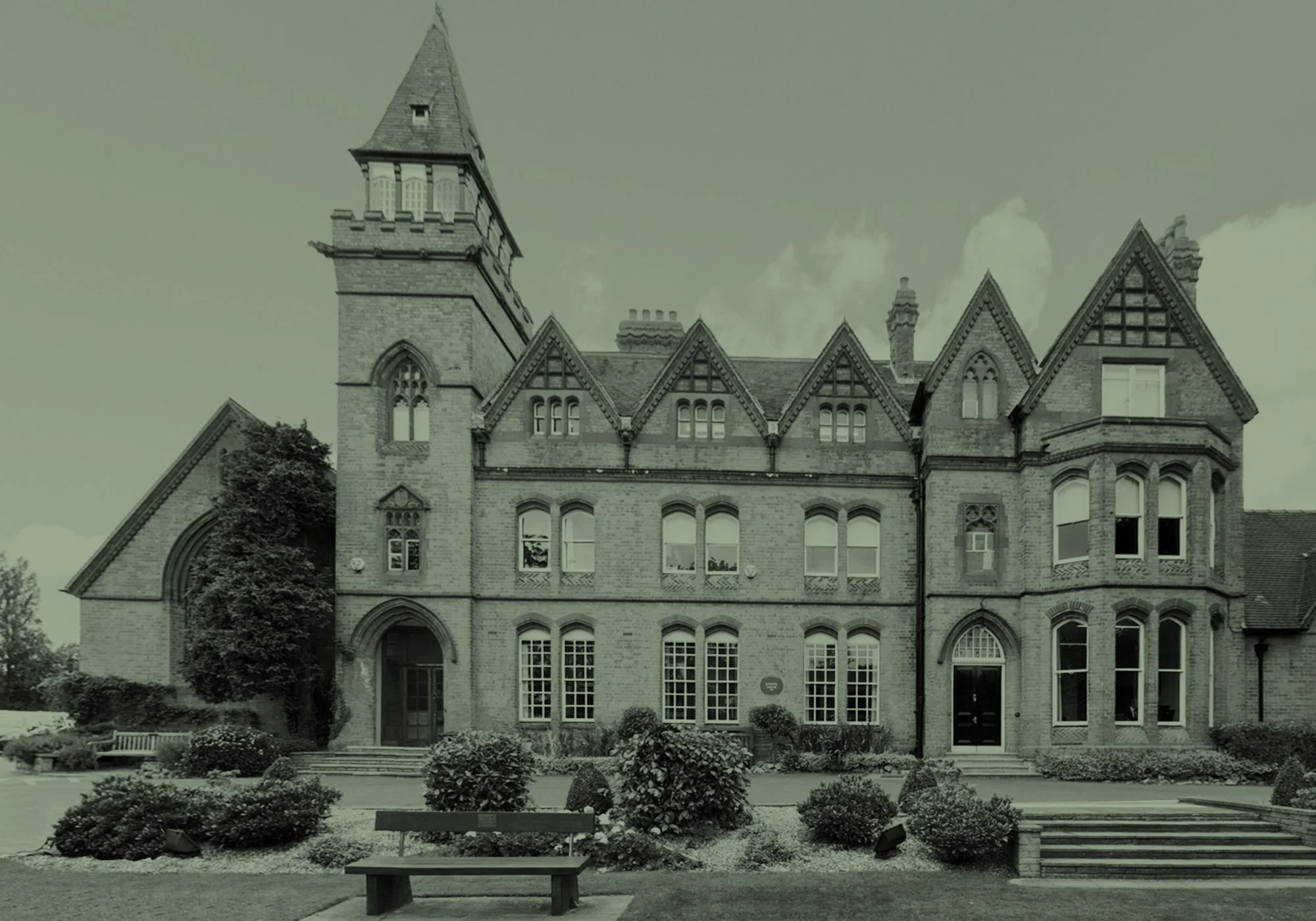 A historic brick building with gothic architectural features, including multiple peaked roofs, arched windows, and a tower, surrounded by well-maintained landscaping with bushes, trees, benches, and a pathway in the foreground.