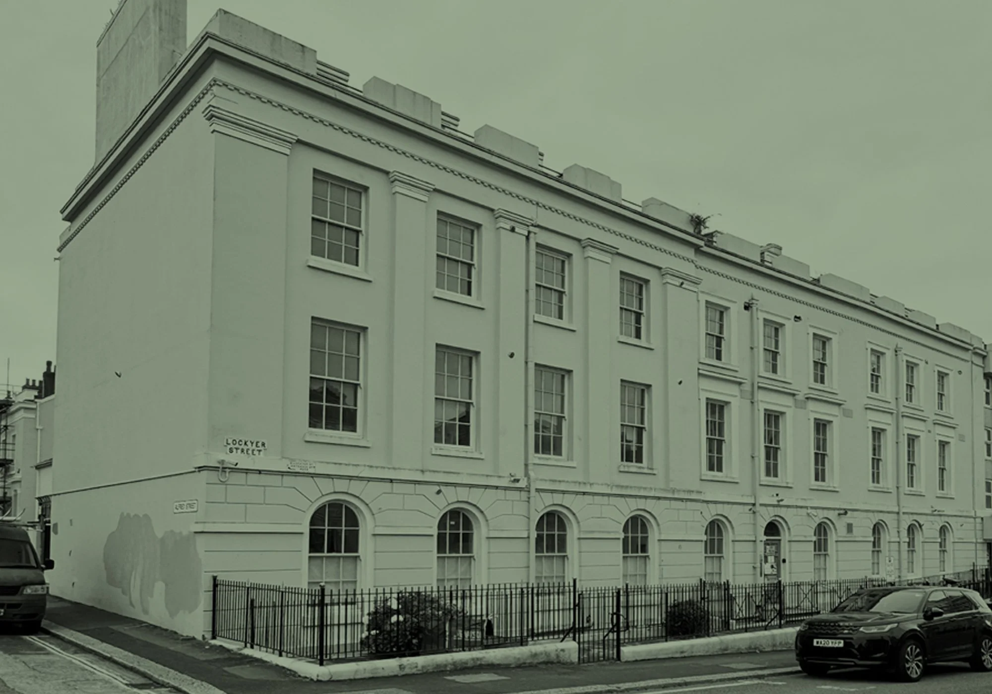 A white multi-story building on a street corner with several windows, a small fenced garden, and parked cars in front.
